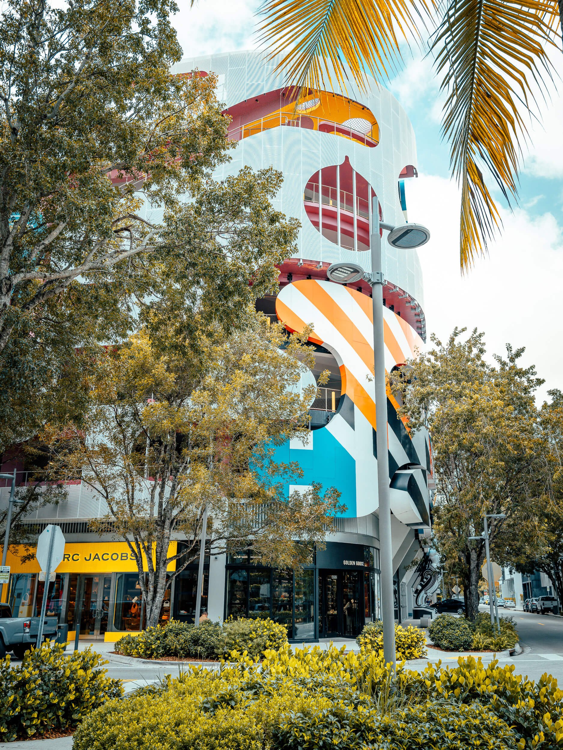 The vibrant façade of the Museum Garage rises behind lush tropical greenery, its bold geometric patterns and sweeping bands of color transforming a parking structure into a striking work of public art. Framed by trees and palms, the scene captures th