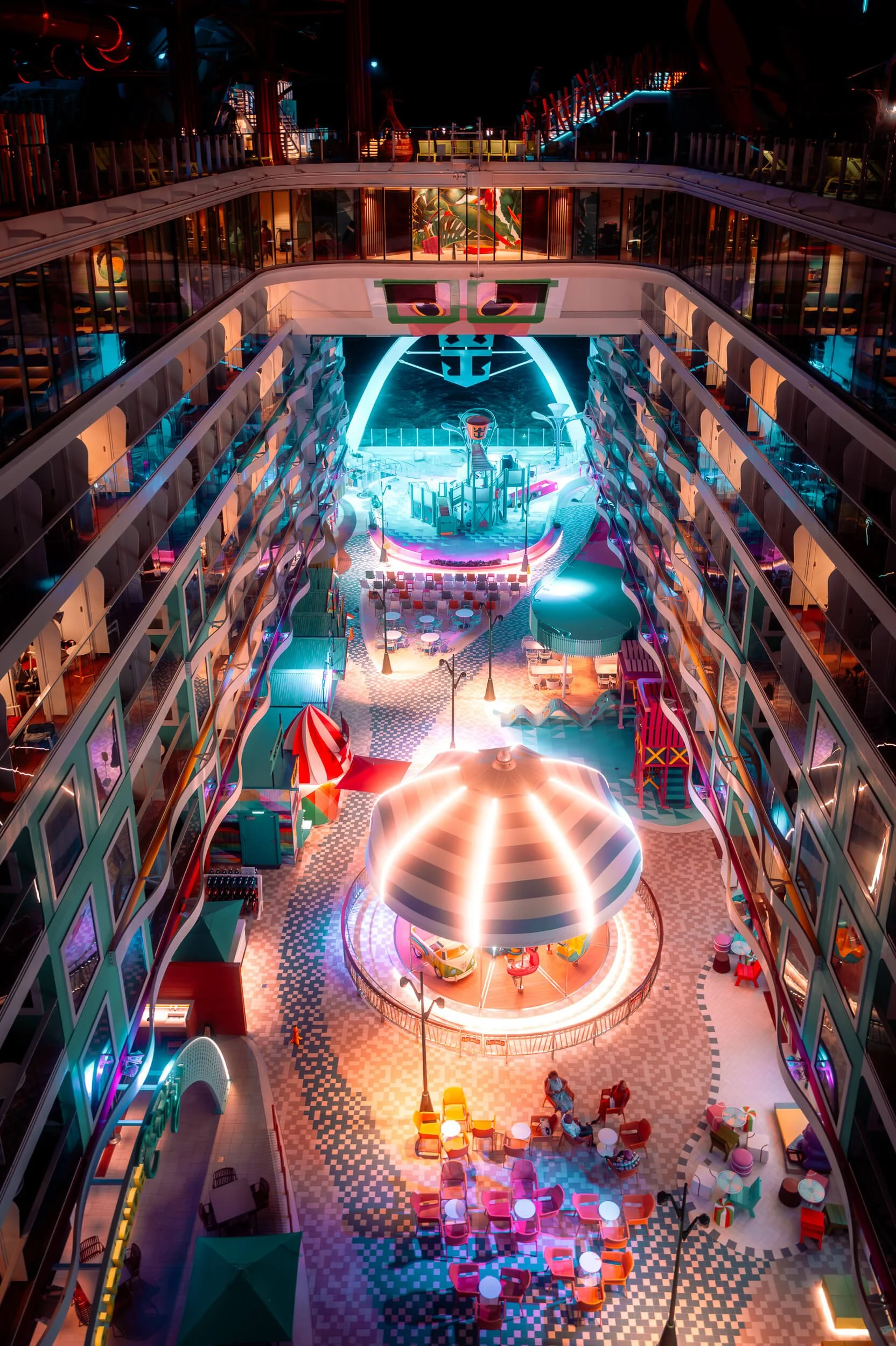 A dazzling nighttime view inside the Icon of the Seas reveals a vibrant open-air atrium alive with color, light, and movement. Neon accents, glowing architectural arches, and playful design elements surround a central carousel, creating a dreamlike s