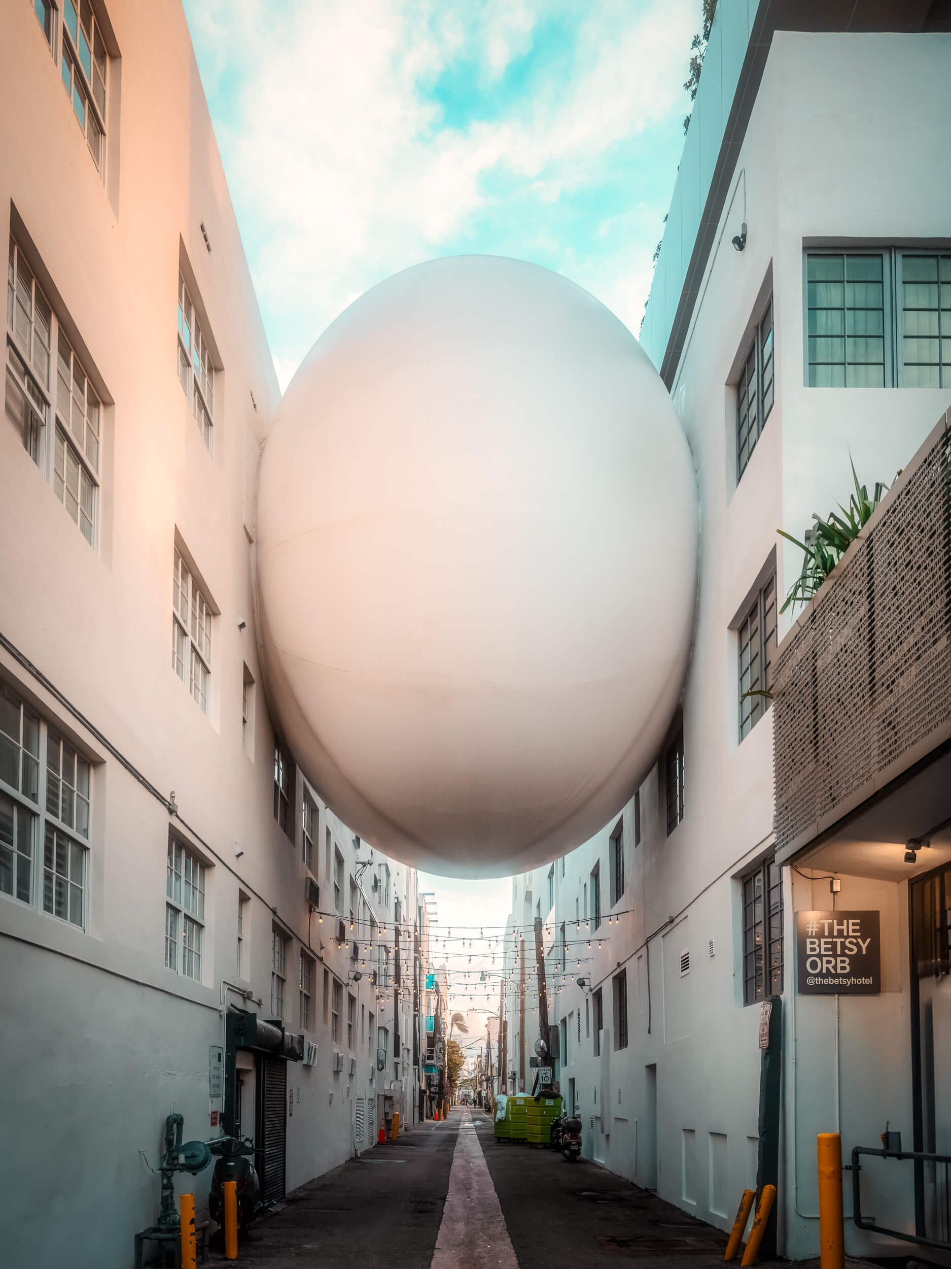 A massive white orb appears suspended between two buildings, transforming a quiet alley into a surreal urban scene. Framed by clean architectural lines and a soft pastel sky, the installation creates a striking contrast between minimal modern design 