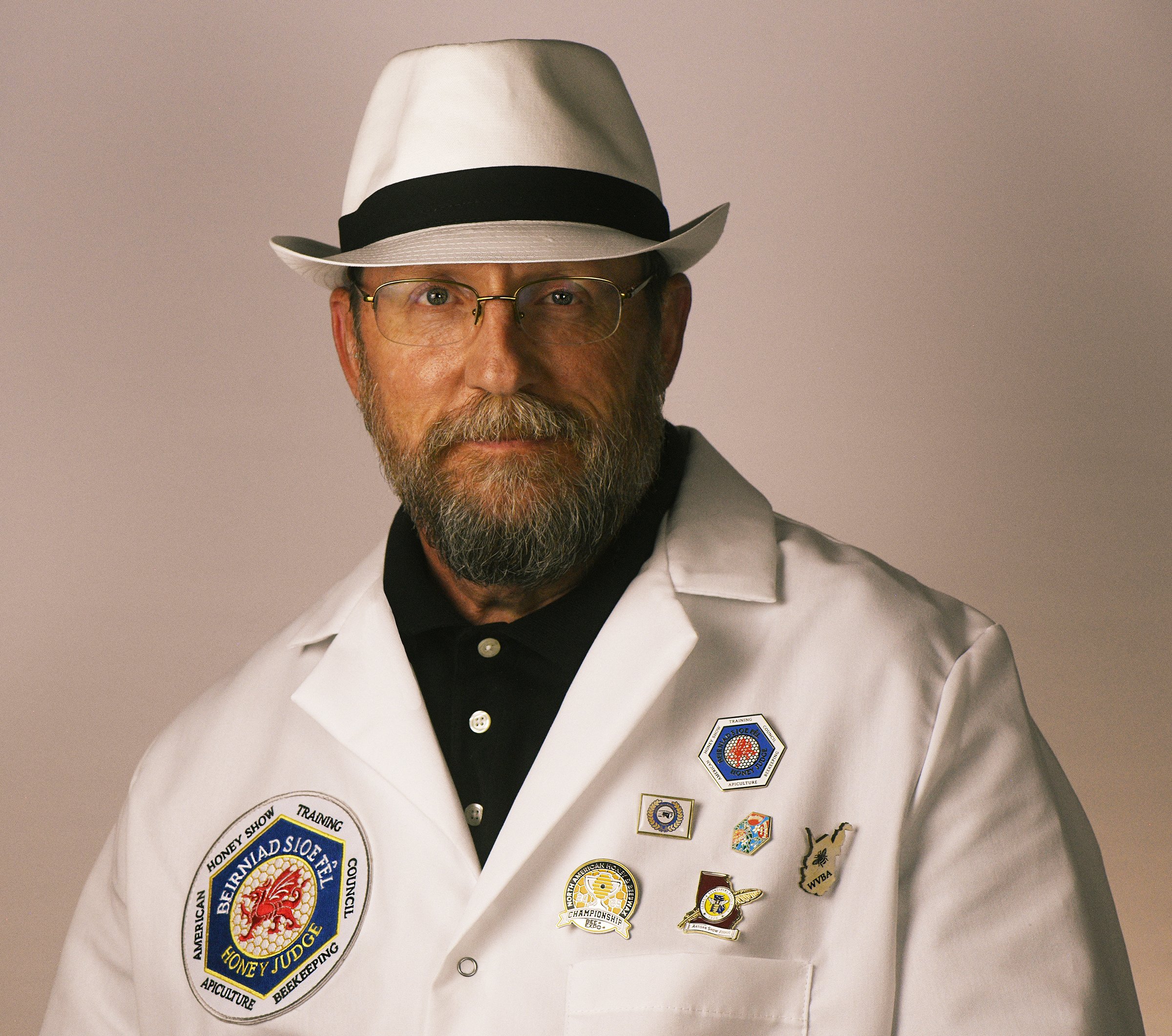 A man with glasses and a beard wearing a white fedora, white lab coat with pins, and a black shirt.