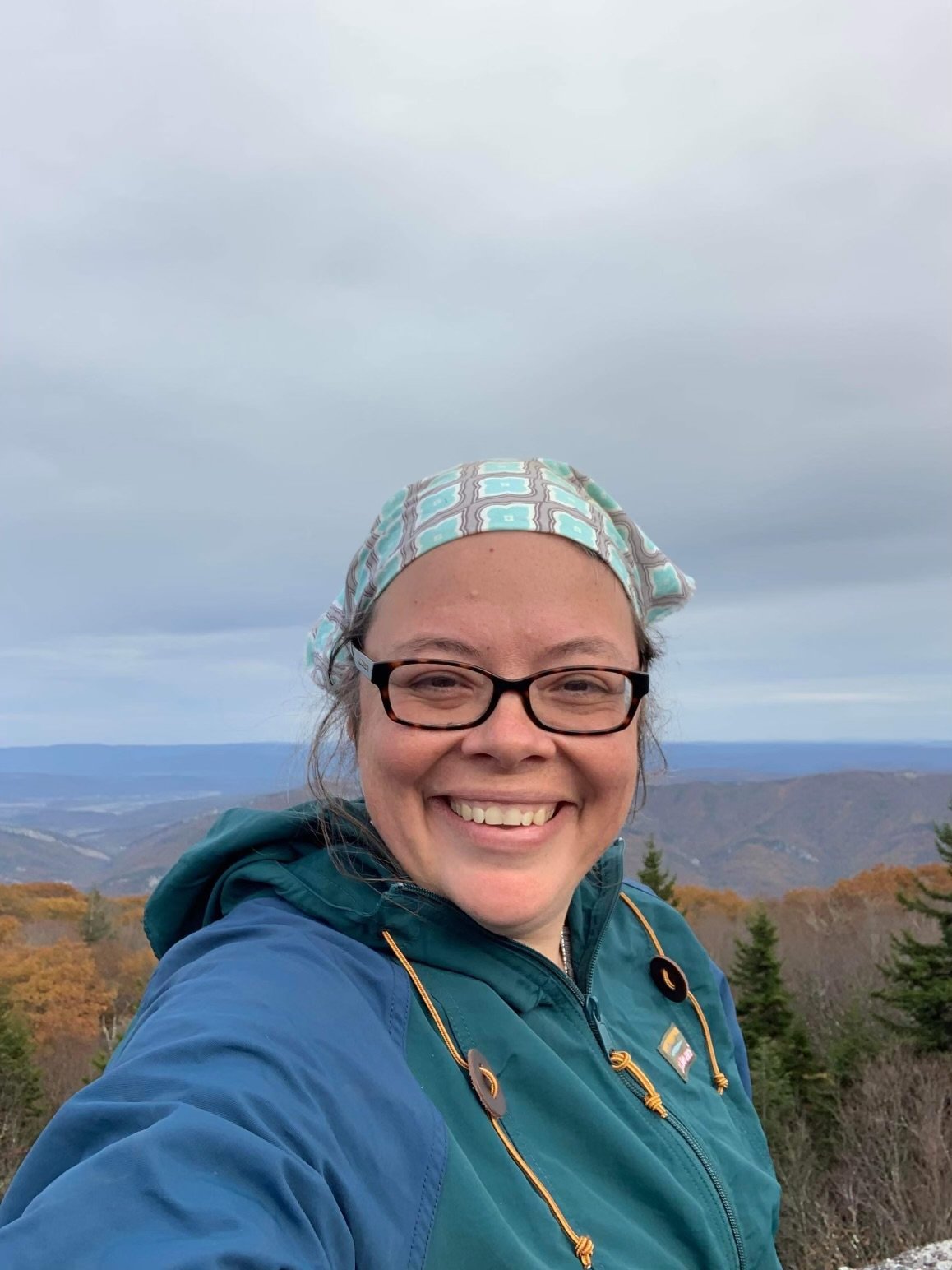 A smiling woman wearing glasses, a light plaid headscarf, and a blue jacket, standing outdoors with a scenic view of rolling hills and a cloudy sky in the background.