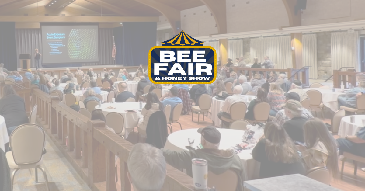 A man is giving a presentation in front of an audience at the Bee Fair Spring Summit. The presentation screen shows images, text, and the words 'Queen Rearing the Easy Way' by Greg Burns. The audience is seated and listening, with some attendees wearing hats and casual clothing. The scene takes place in a room with wooden walls and a large logo for the Bee Fair Spring Summit is superimposed in the middle of the image.