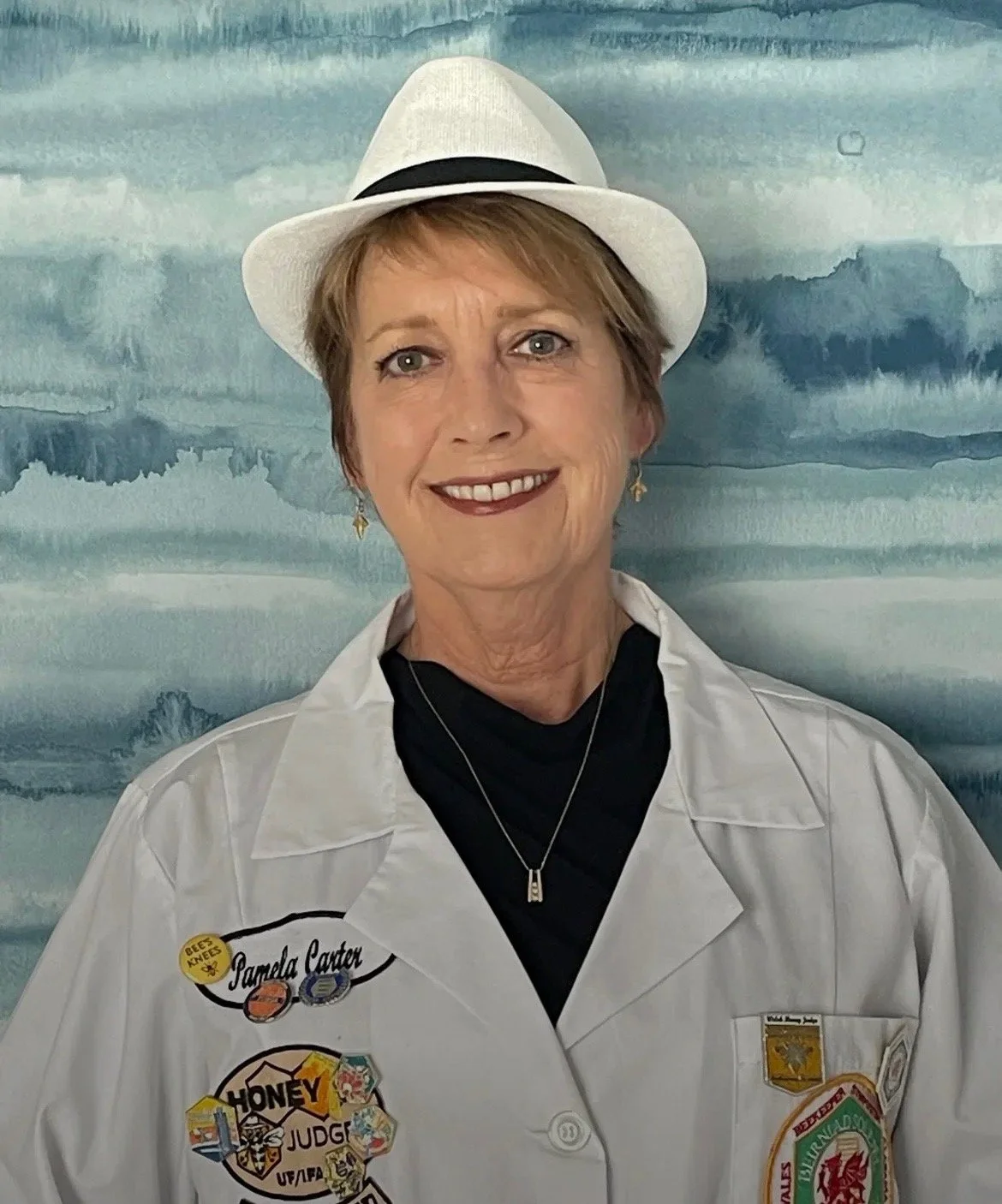 A woman smiling, wearing a white fedora hat, a white jacket with various patches, and a black shirt underneath, standing in front of a watercolor-style blue and white background.