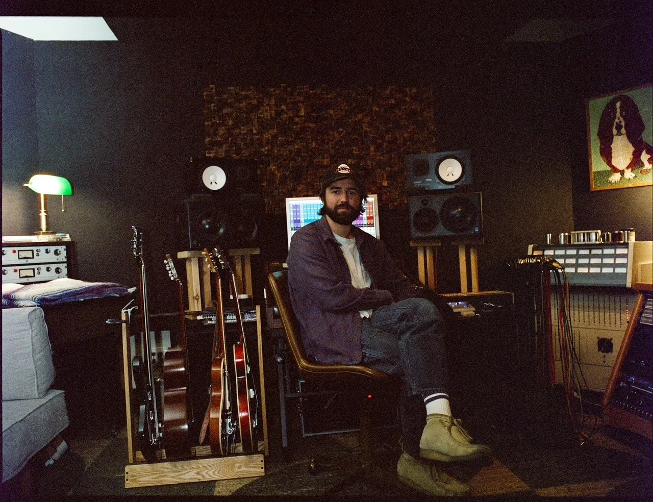 Man with a beard sitting in a music studio surrounded by guitars, audio equipment, and speakers, with a computer monitor displaying audio tracks in the background.
