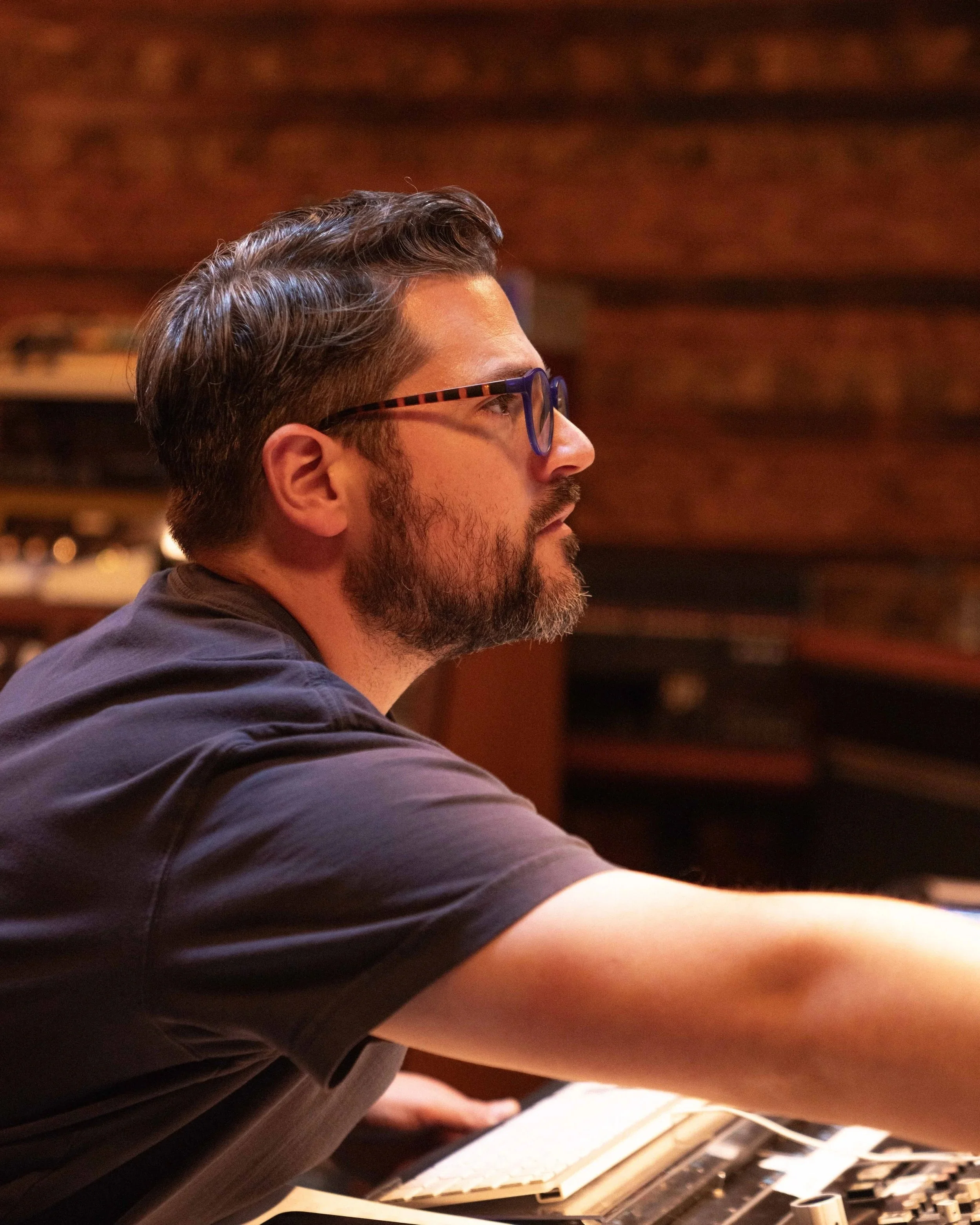Profile of a man with dark hair, glasses, and facial hair, wearing a gray shirt, sitting in a dimly lit room with wood accents.