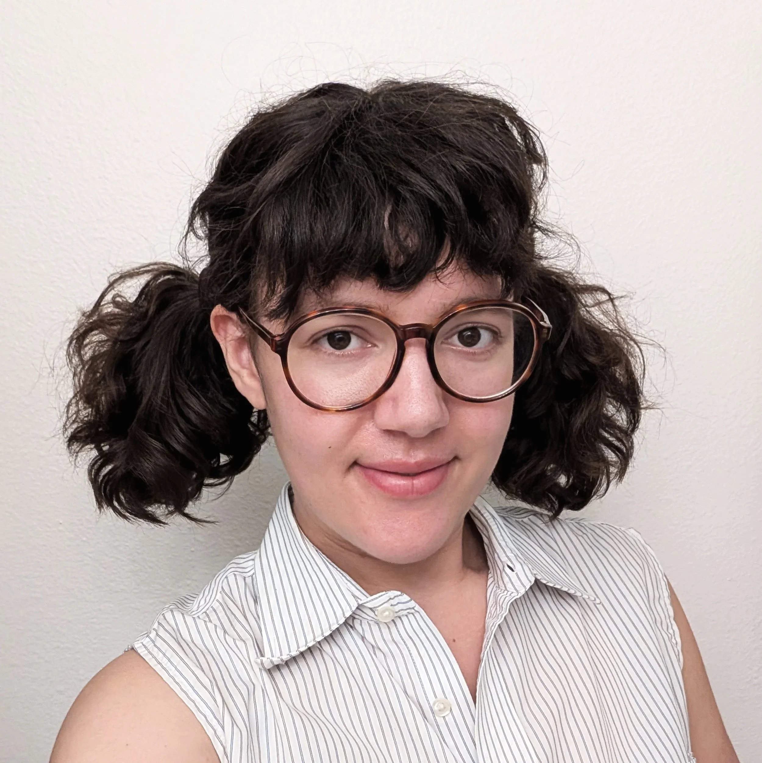 Close-up of woman with curly brown hair, wearing glasses with round frames, white and gray striped sleeveless shirt, standing against a plain white wall.