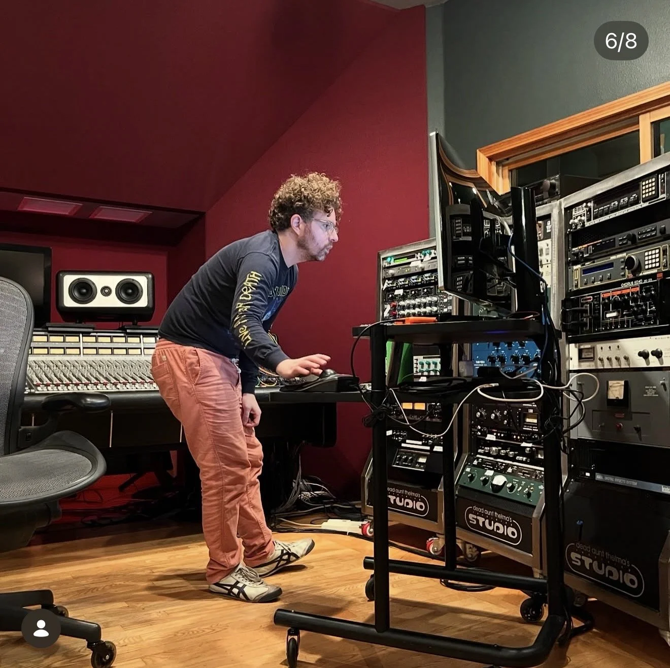 A man with curly hair and glasses is operating audio equipment in a music studio with red and gray walls. He is leaning over a mixer and standing next to rack-mounted audio gear on wheels, with studio monitors and a chair visible.