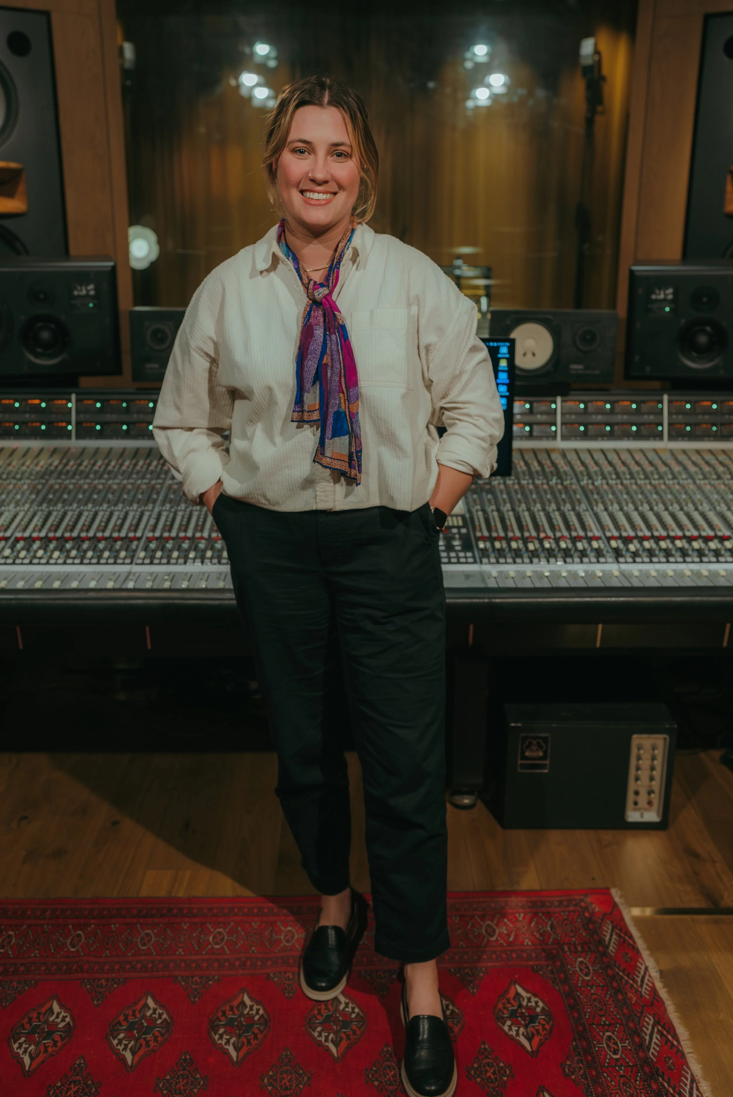 Woman in a recording studio surrounded by audio equipment, wearing a white shirt, colorful scarf, black pants, and black shoes.