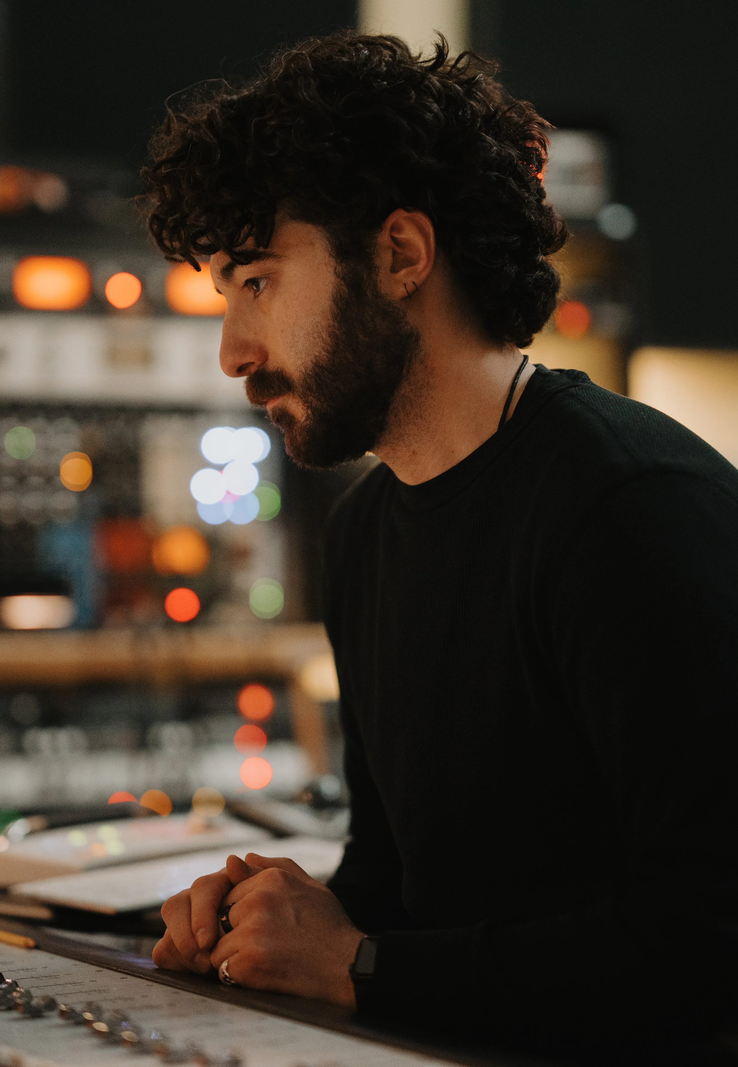 Side profile of a man with curly hair and a beard, wearing a black shirt and an earring, leaning on a table in a dimly lit indoor space with blurred colorful lights in the background.