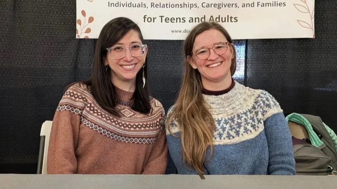 Two people sitting together at a table, smiling, wearing sweaters.