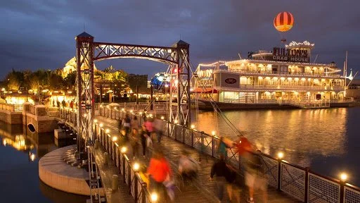 Bridge leading to a lit-up waterfront area with a large paddle steamer and hot air balloon.