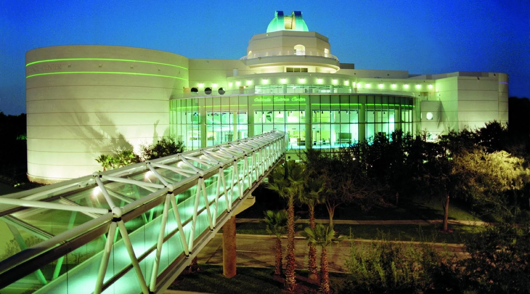Orlando Science Center at night, lit in green with a glass bridge.