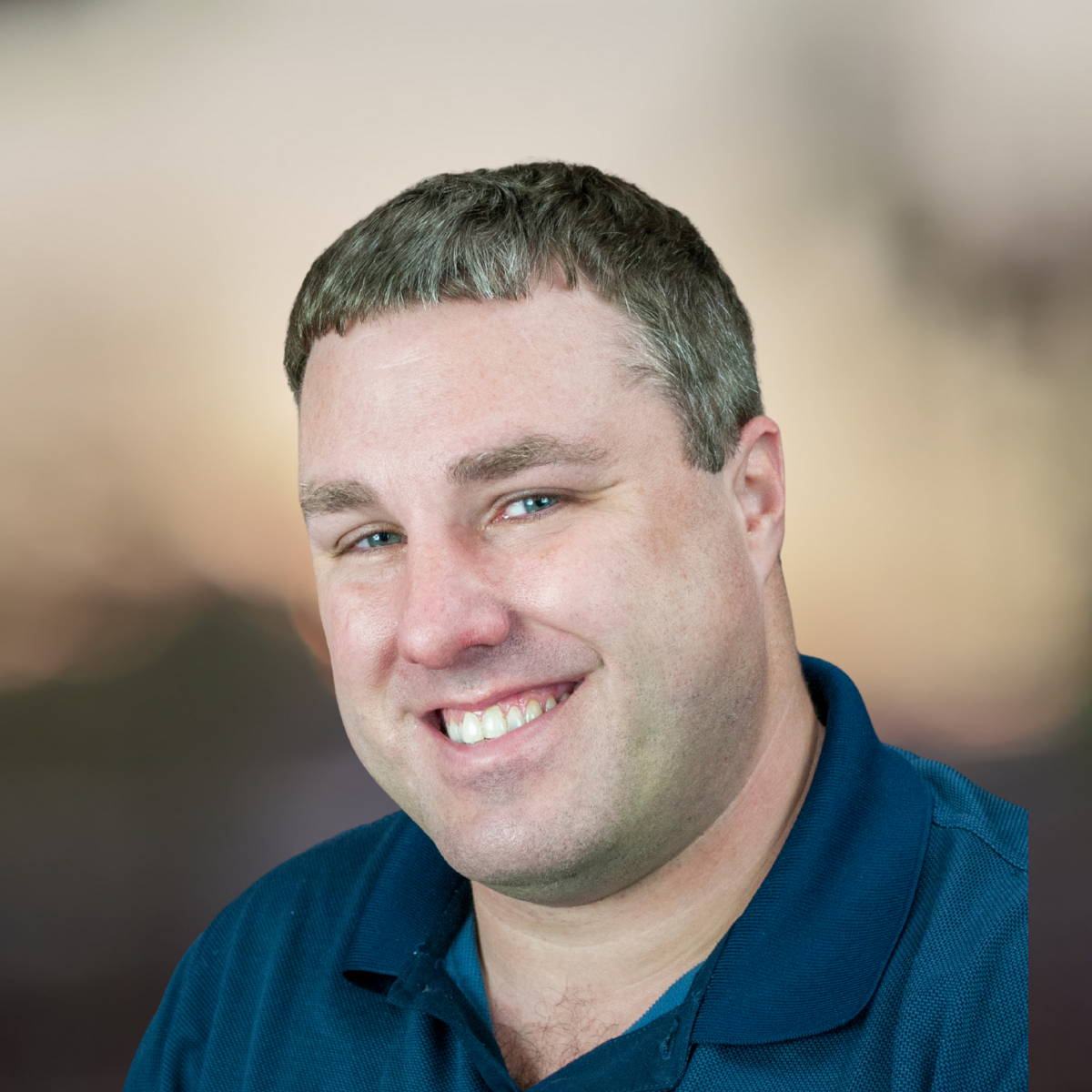 Man in a blue polo shirt smiling against a brown background.