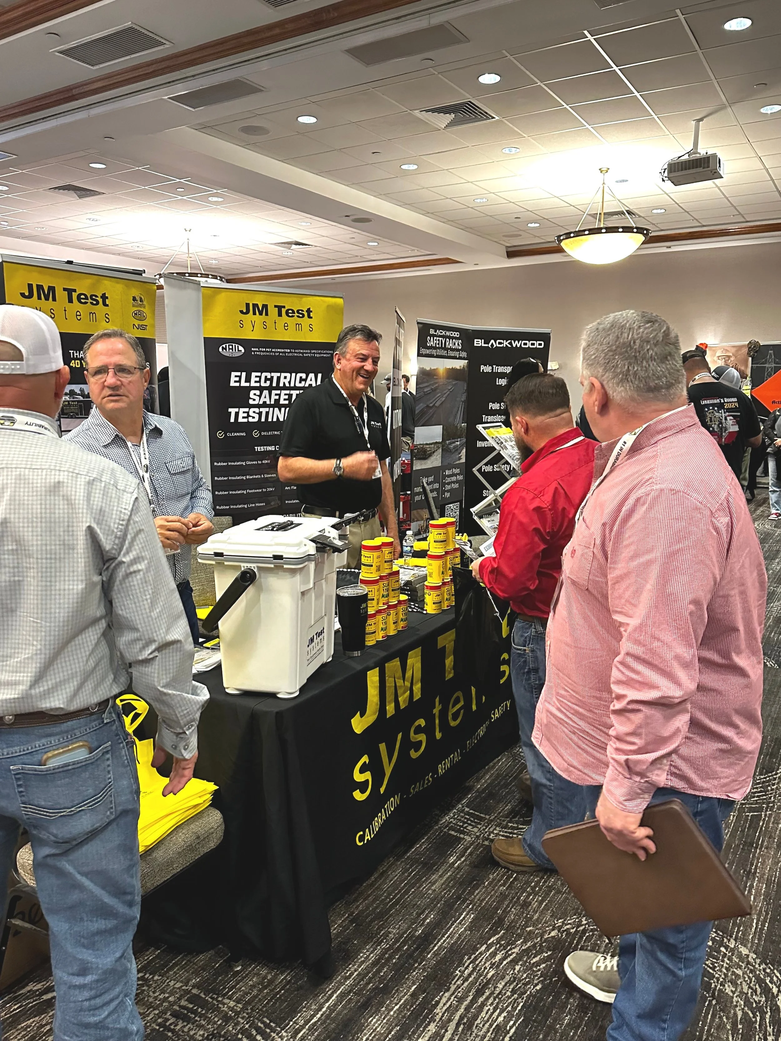 JM Test Systems display, featuring products and informational banners, surrounded by attendees at the 2025 Trainers Conference and Tradeshow