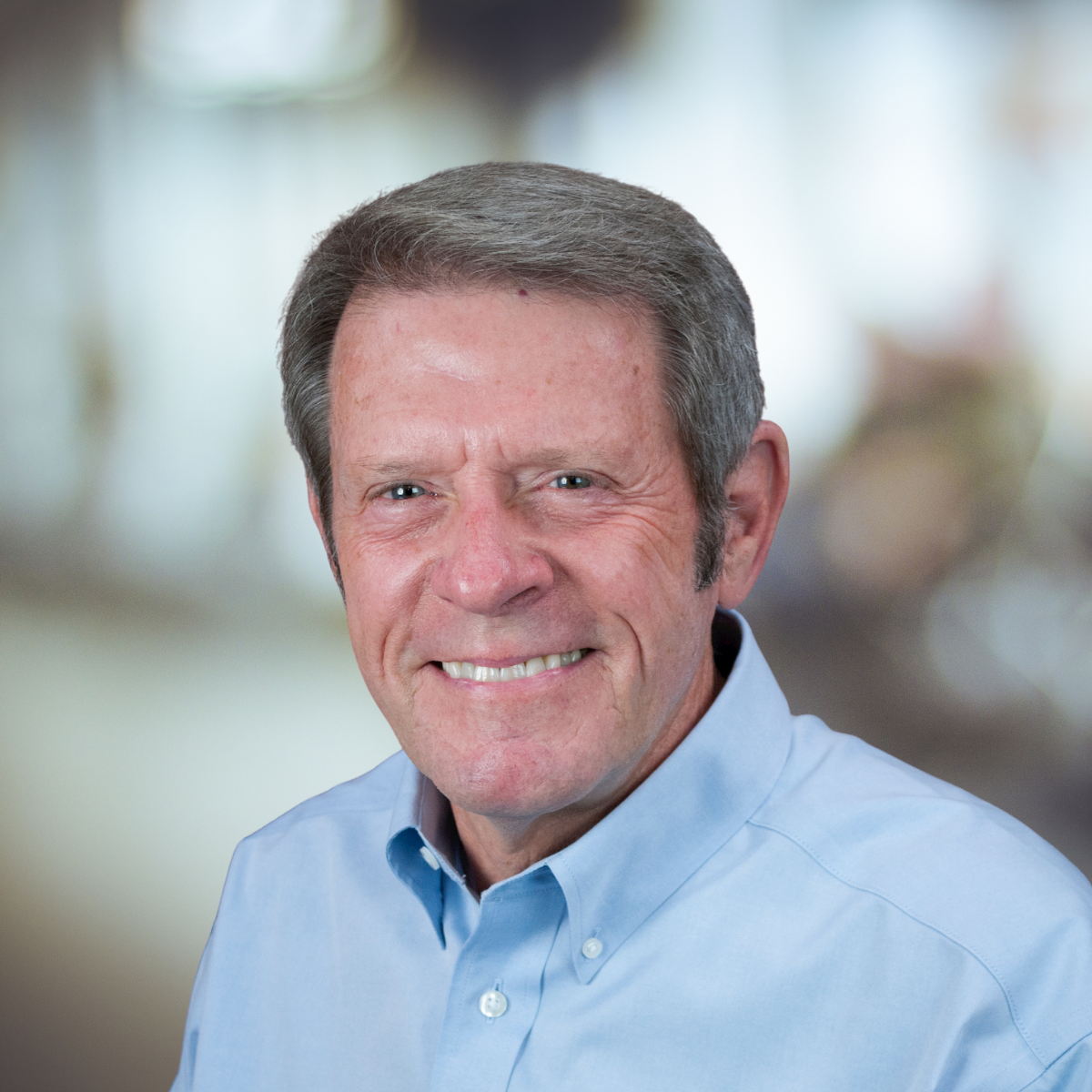 Man wearing a light blue shirt with "Safety Management International" logo, smiling, with a brown textured background.