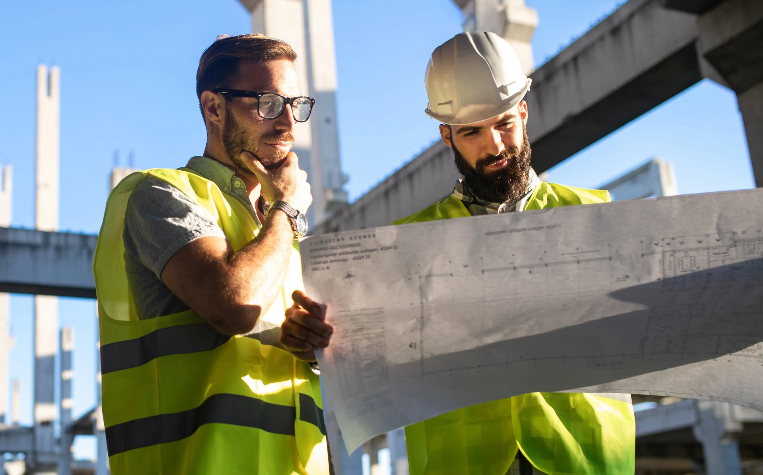 Two construction workers in yellow safety vests and helmets review blueprints at a construction site with steel structures and a blue sky.