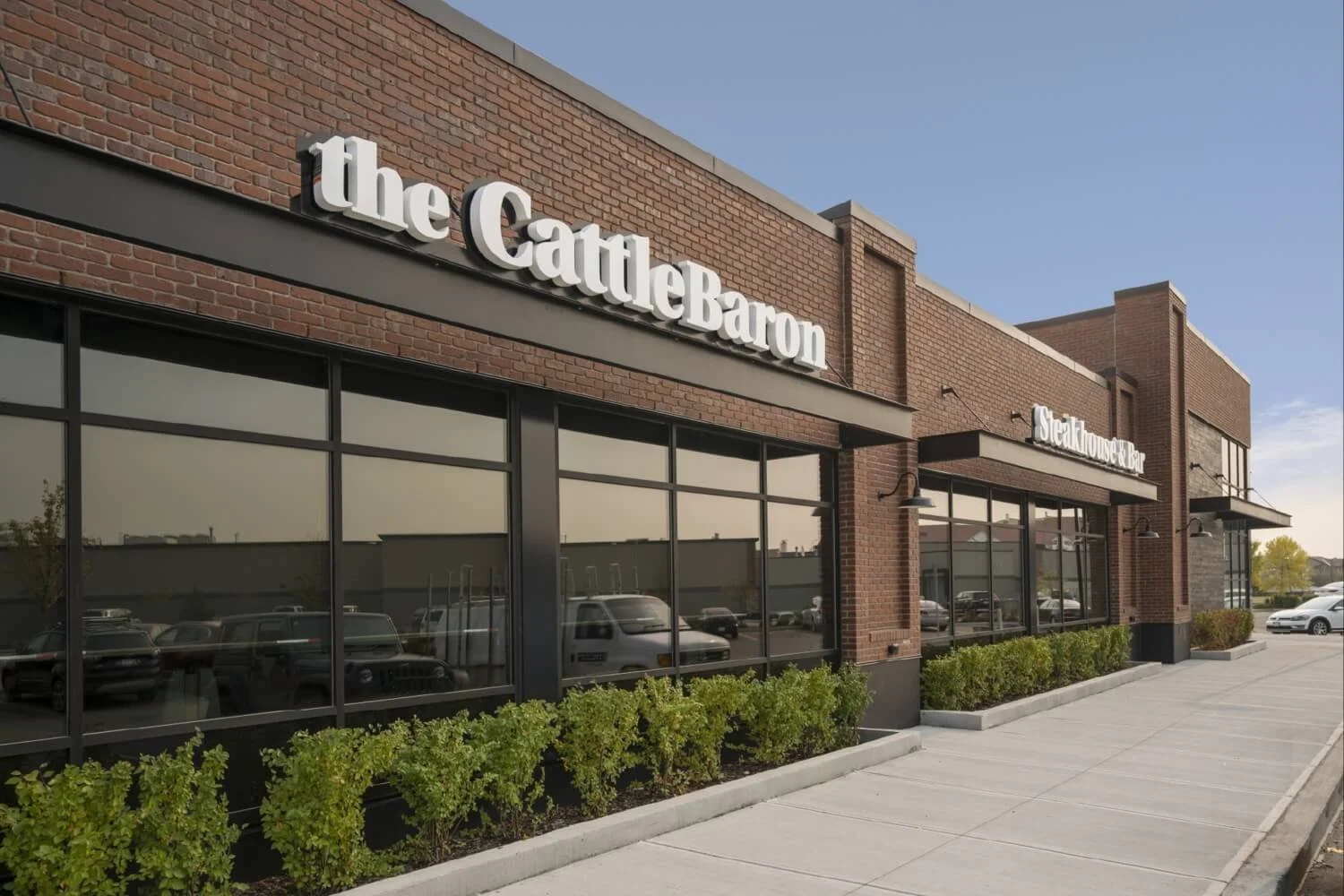 Exterior of the CattleBaron restaurant with brick facade, large windows, and green shrubs in front, under a blue sky.