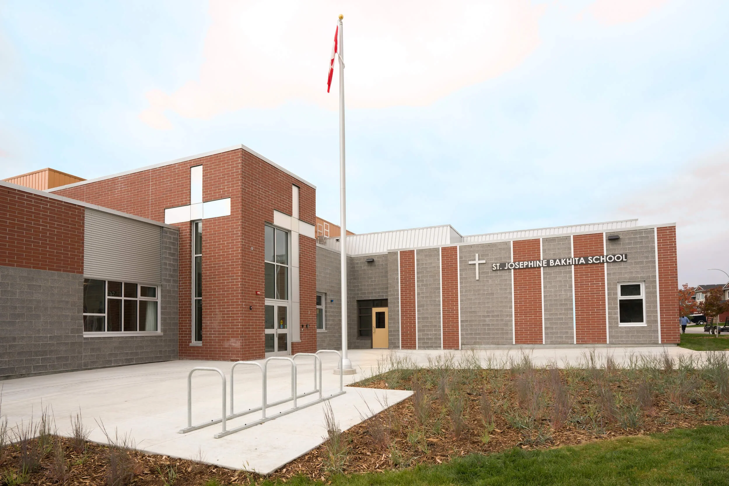 Exterior of St. Josephine Bakhita School with brick and gray wall construction, a flagpole with a flag, and a grassy area.