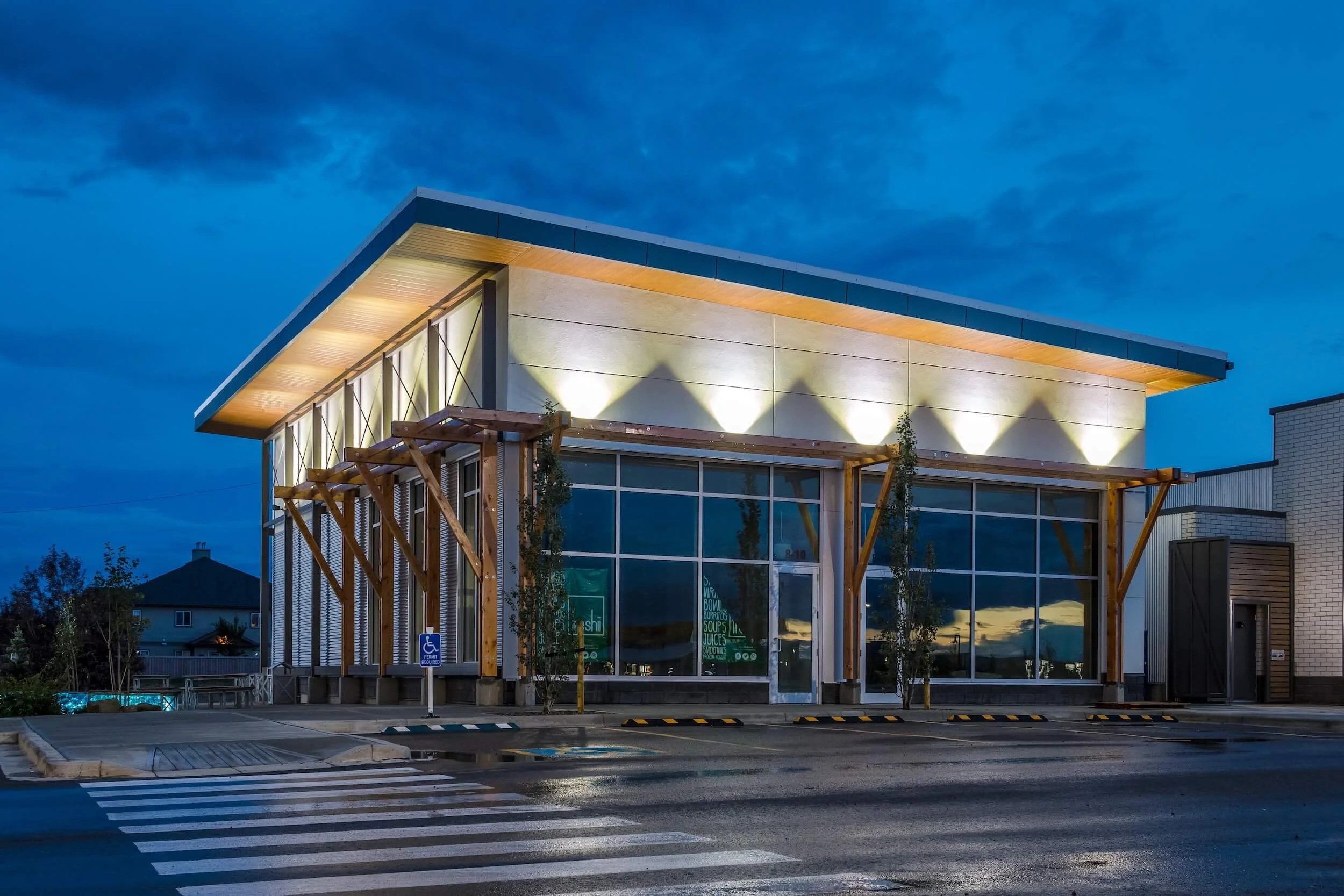 Modern building with large glass windows, wooden supports, and exterior lighting at dusk, in a parking lot with a crosswalk.