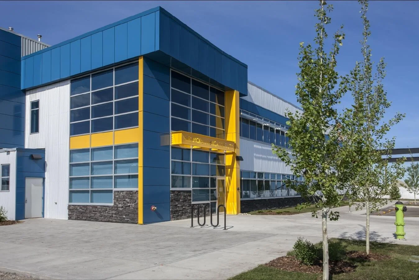 Glenmore Fabricators industrial building with blue, white, and yellow exterior, large glass windows, and a small greenery area with trees in the foreground.