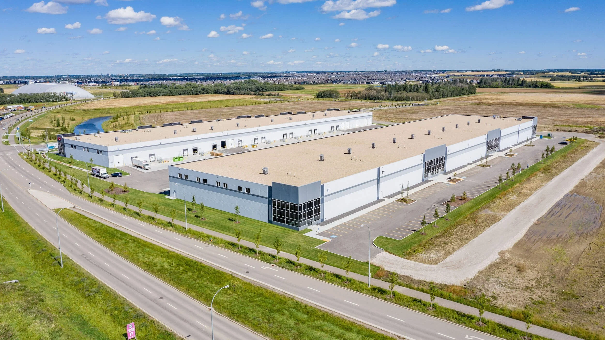 A large, modern industrial building with a parking lot, situated in a semi-rural area with open fields and a small waterway nearby.