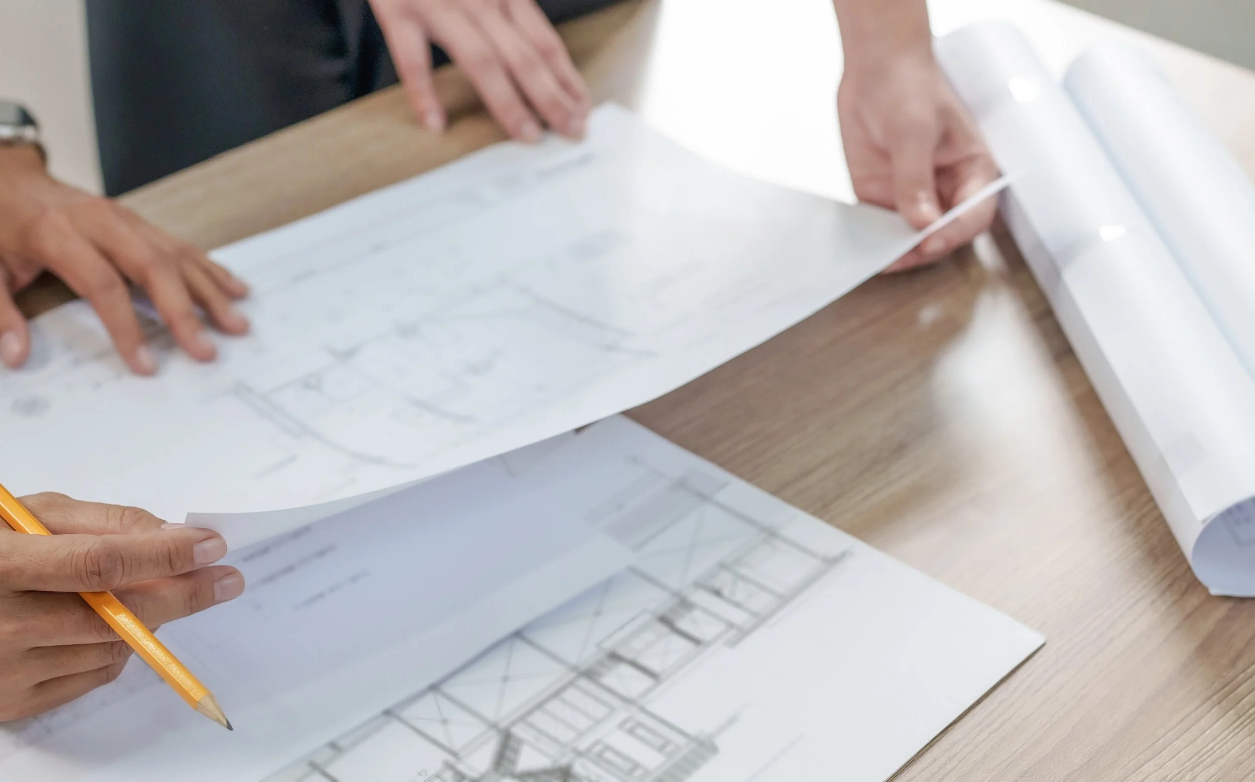 Two people reviewing architectural blueprints on a desk, one holding a pencil and the other flipping through the plans.