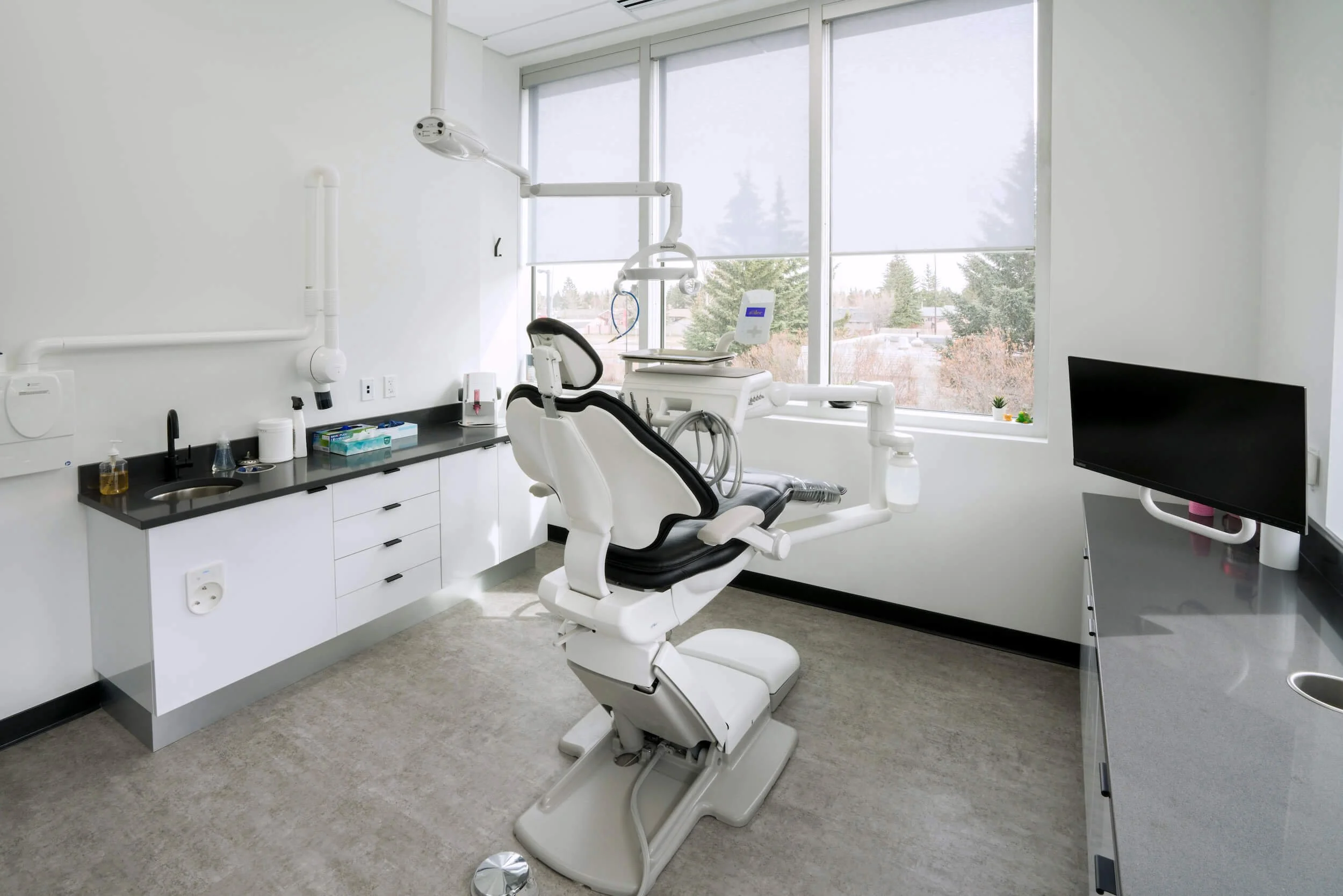 Dental examination room with a dental chair, computer monitor, and white cabinets with dental supplies. Large windows with blinds letting in natural light.