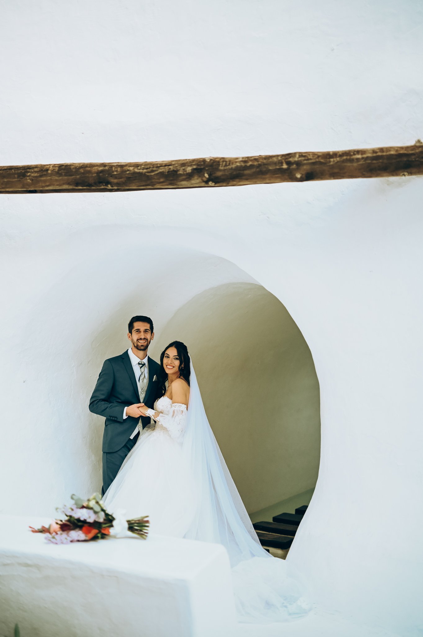A couple in wedding attire posing in front of a white curved architectural background, with a bouquet of flowers on a table in the foreground.