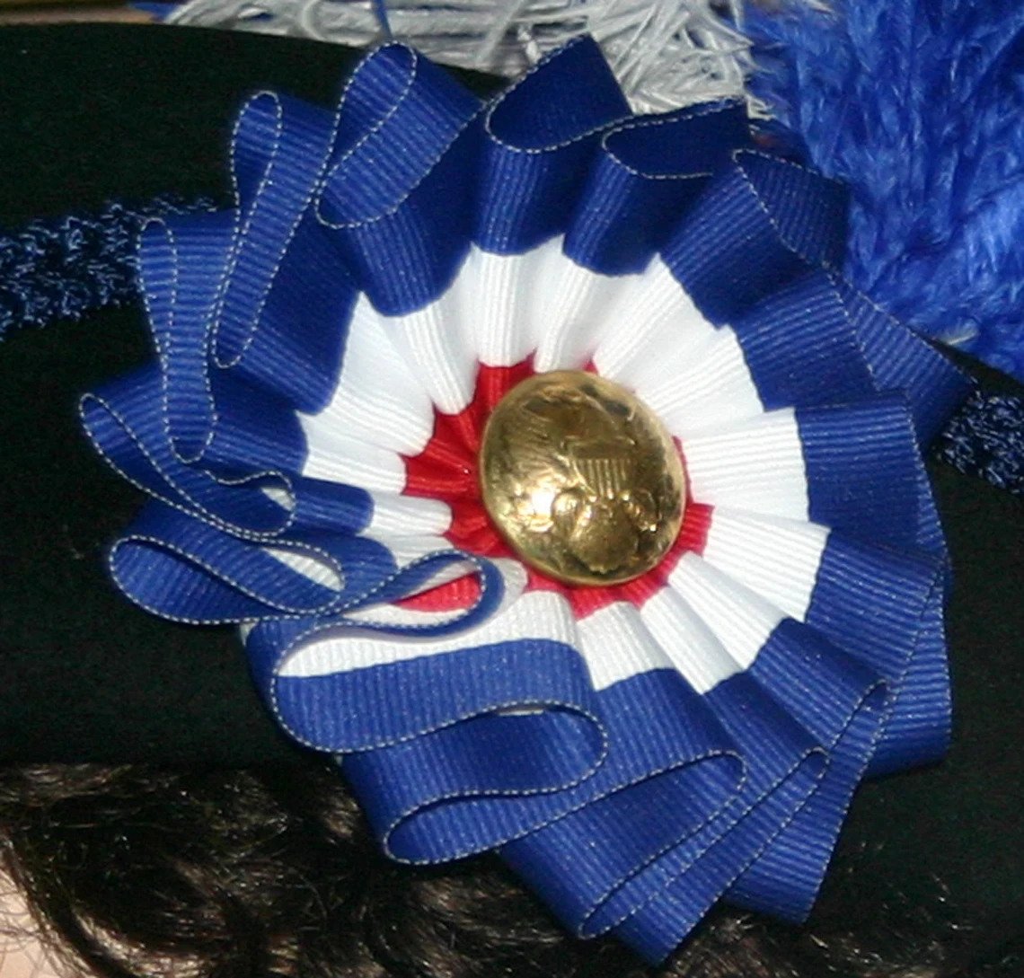 Detailed close-up of red, white, and blue cockade with brass Civil War buttons on navy blue wool hat.