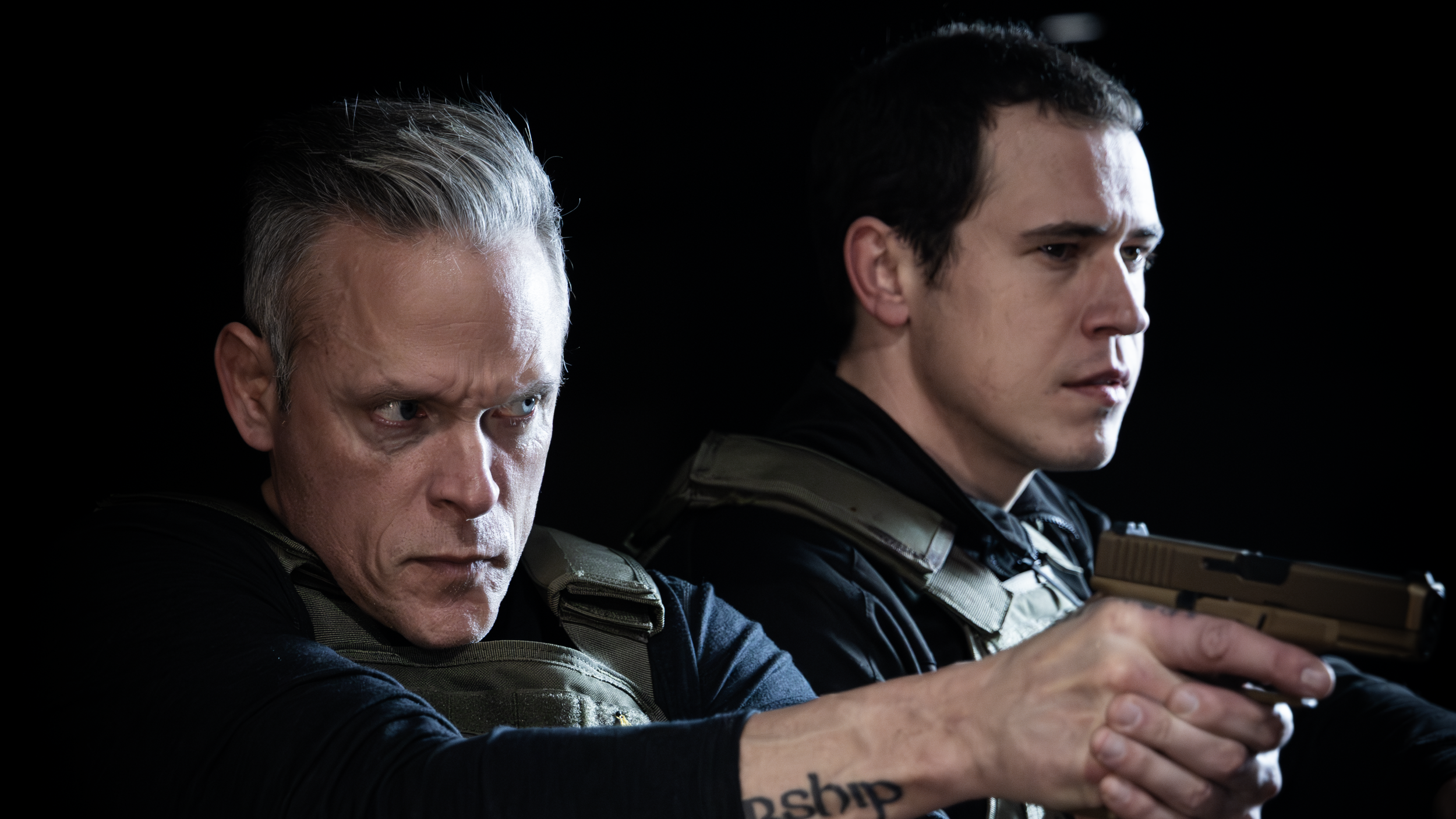 Two men with serious expressions, aiming guns, against a black background. They are wearing tactical gear.