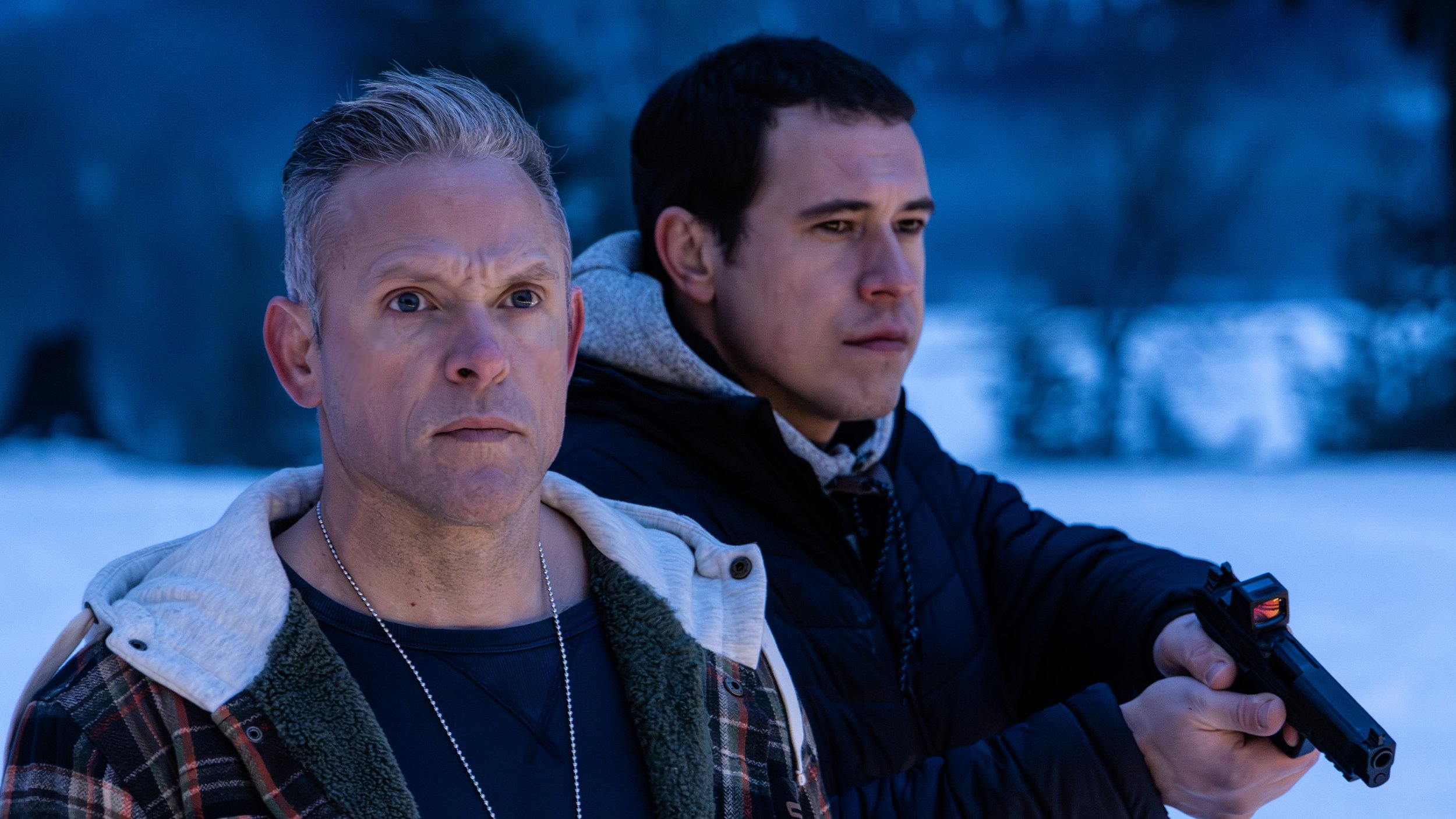 Two men standing outside in a cold environment during dusk or dawn, one is older with short gray hair, wearing a plaid jacket and a hoodie, and the other is younger with dark hair, holding a firearm, with a serious expression.