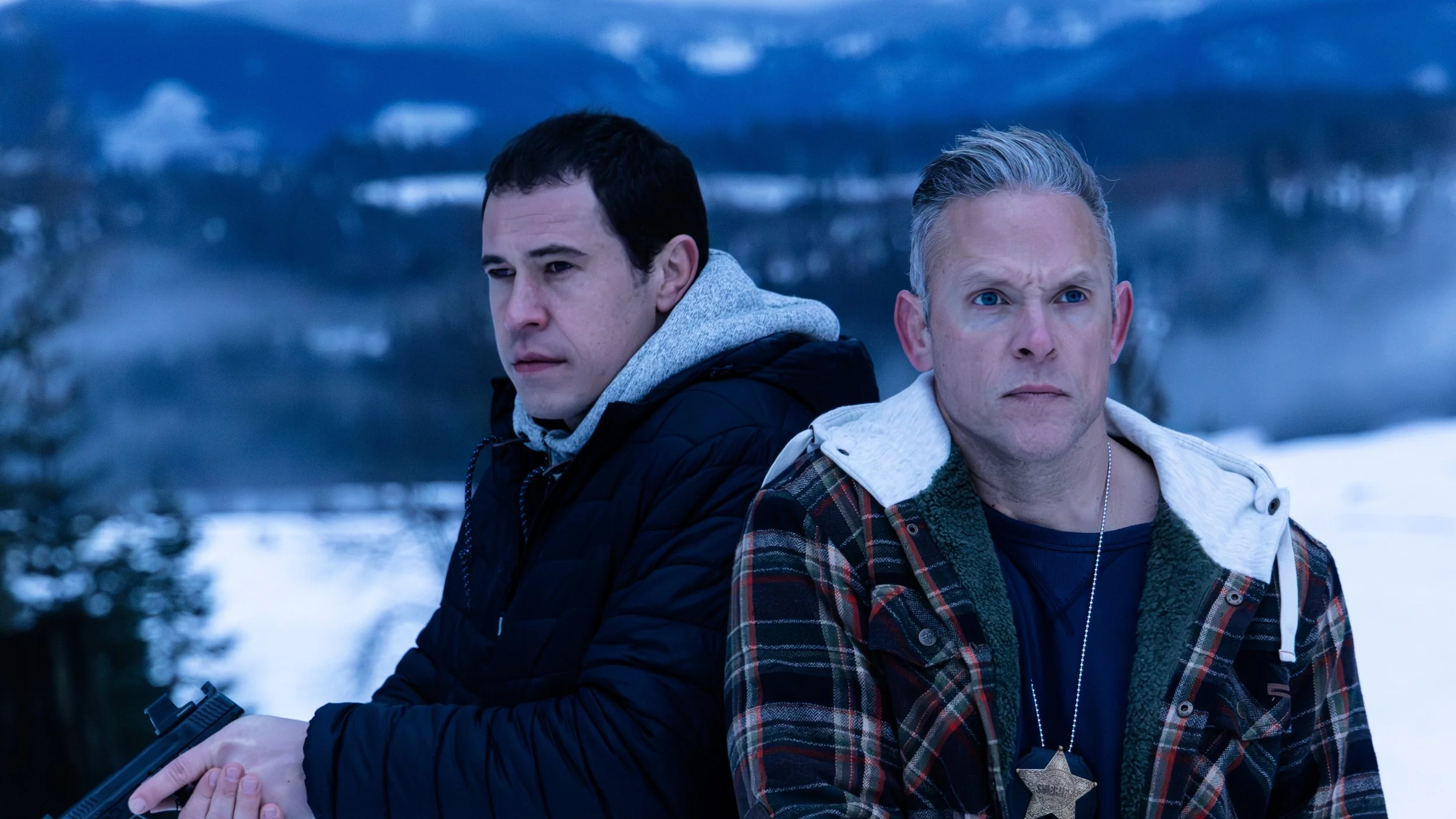 Two men standing outdoors in a snowy landscape with mountains in the background, one holding a gun.