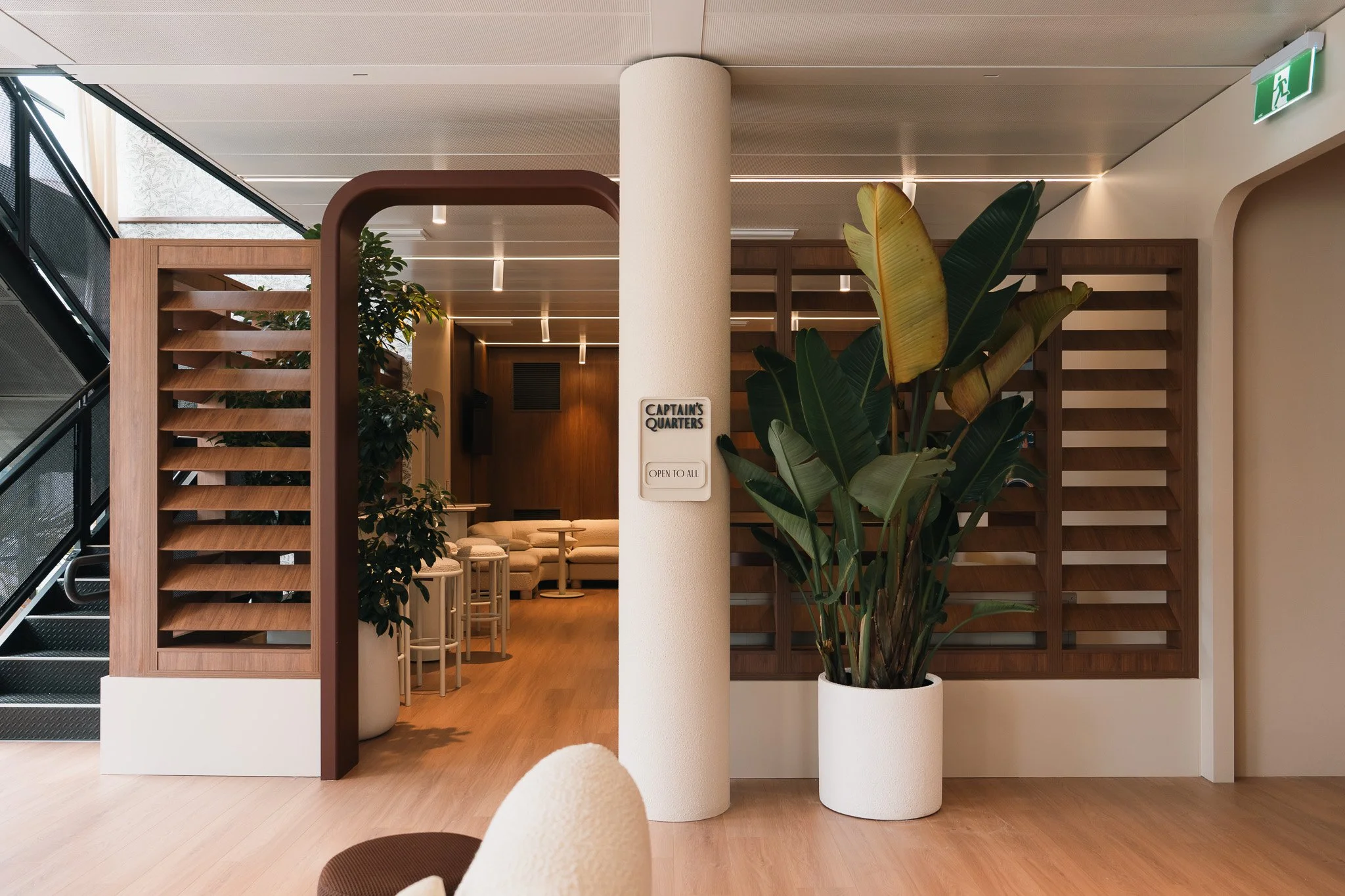 Interior of a modern space with a white pillar, large green potted plant, wooden partitions, furniture, and an emergency exit sign at the ceiling.