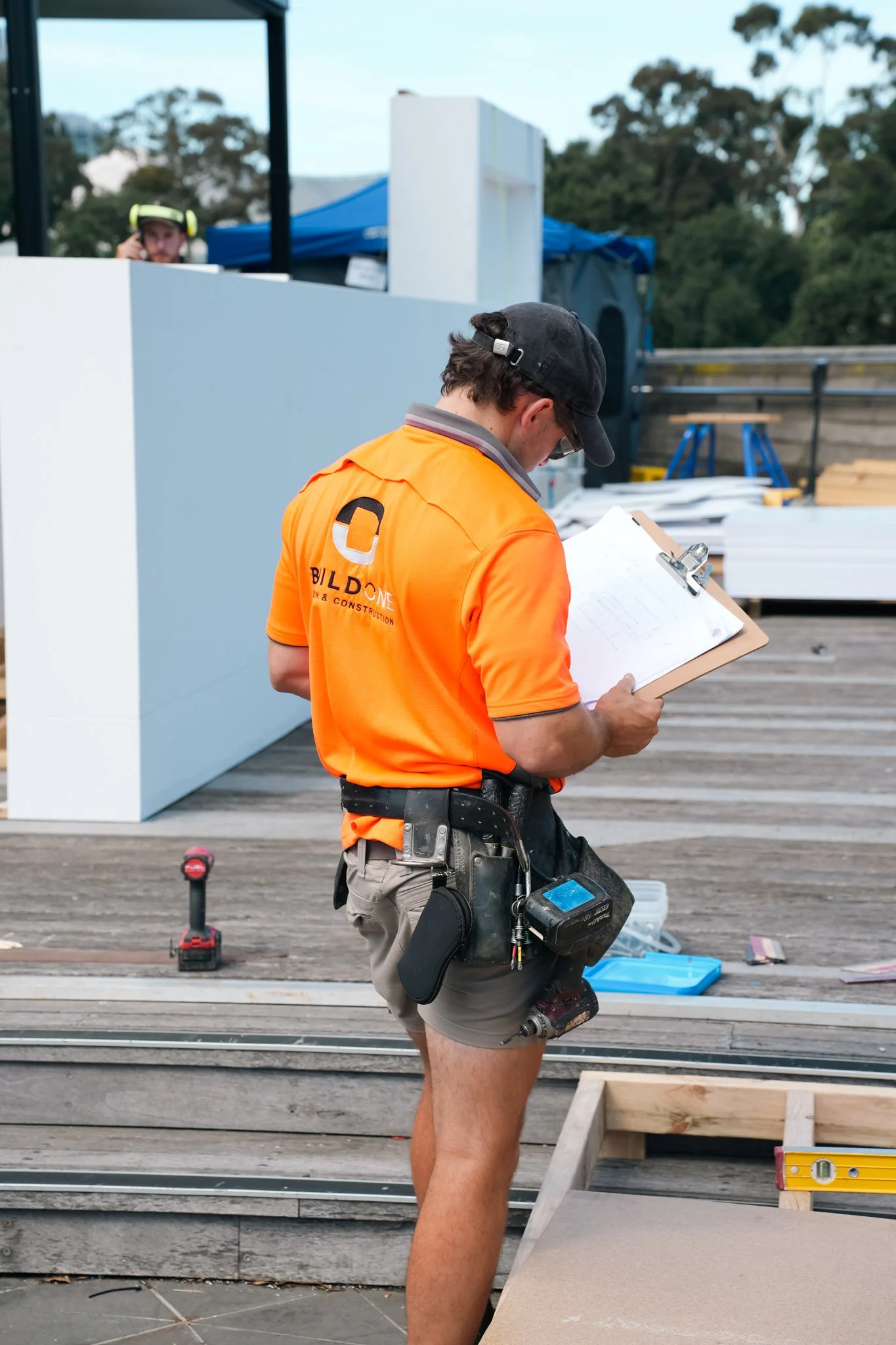 Construction worker in orange shirt, shorts, and black cap holding a clipboard, standing on a wooden deck with construction tools and materials nearby.