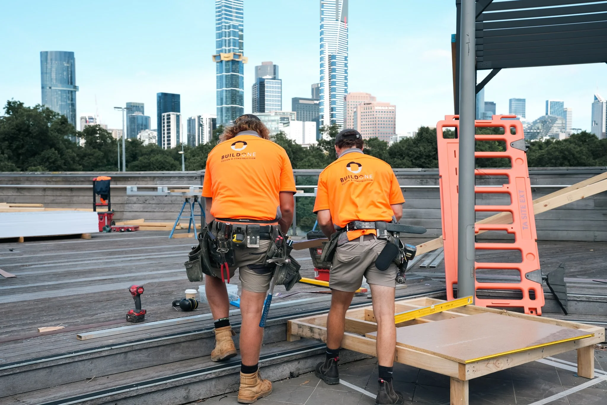 Two construction workers wearing orange shirts and construction harnesses working on a rooftop with city skyline in the background. One worker is wearing brown boots, and both are facing away from the camera, involved in a construction project with w