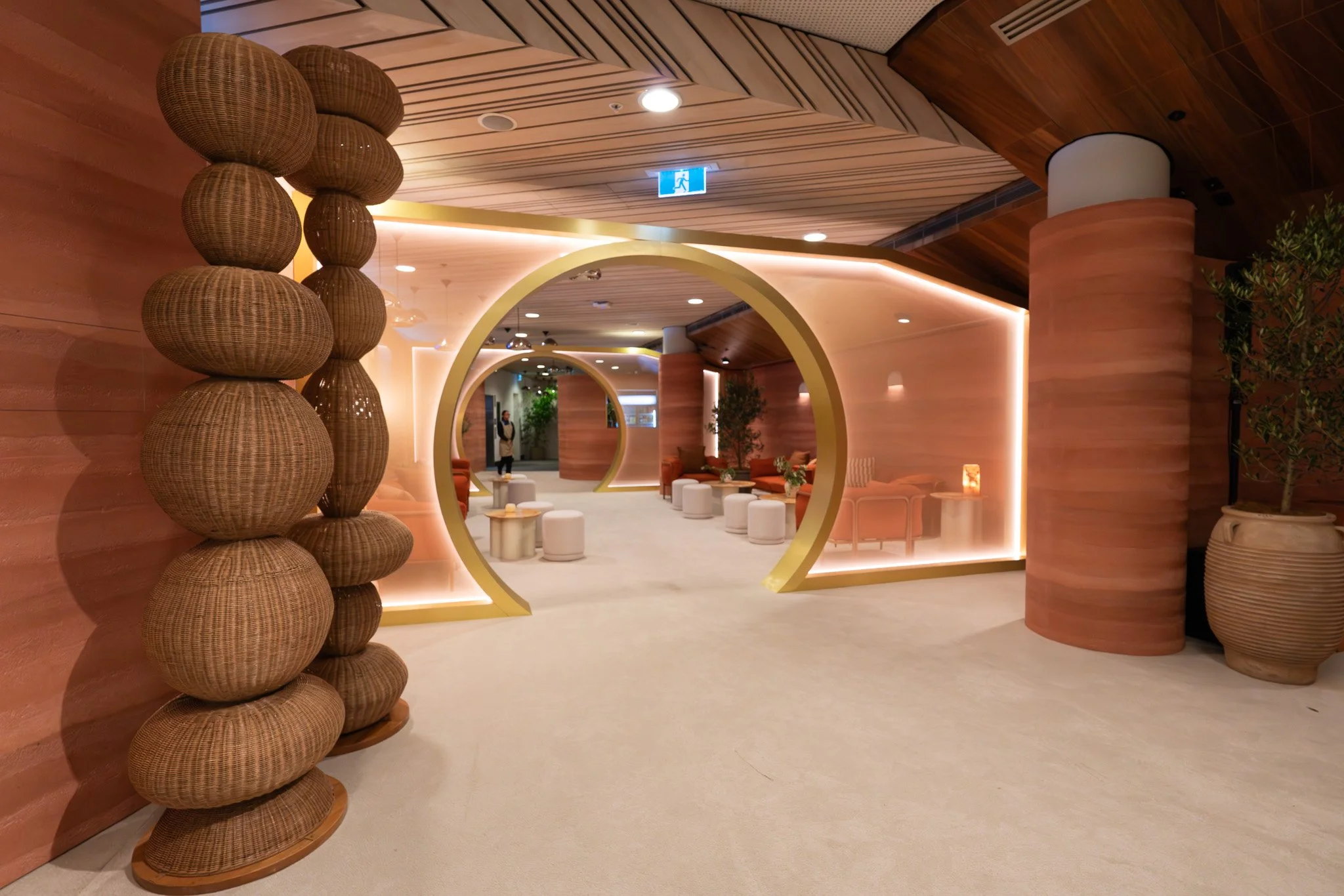 A modern interior space with wooden paneling, decorative circular structures, and lounge seating, illuminated by soft lighting and an emergency exit sign.