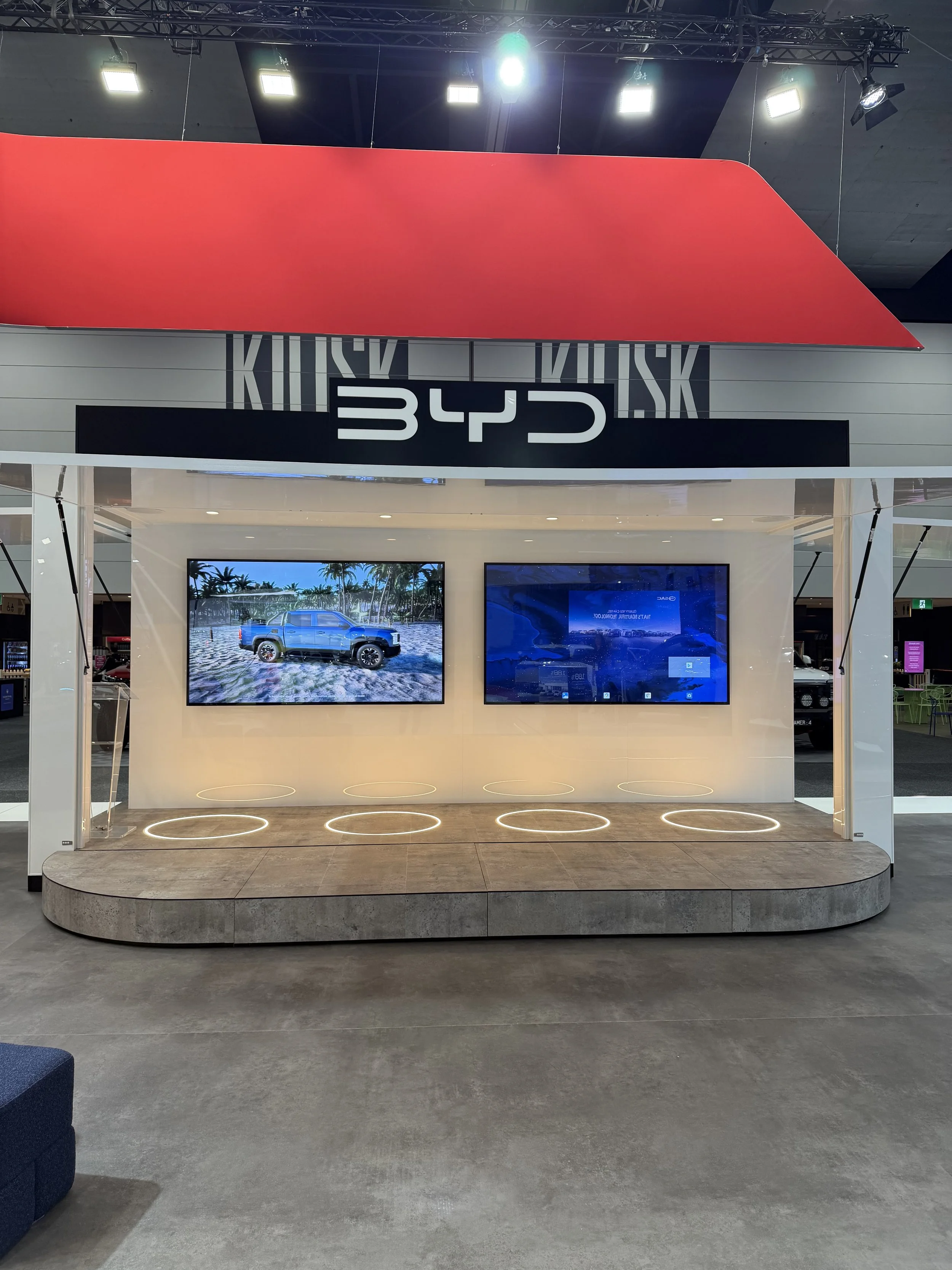 Indoor display area featuring two high-resolution screens showing outdoor scenes with a blue truck and ocean views, with the letters 'BDS' in large black and white sign above, and a red angular canopy overhead. The area has a minimalist design with a