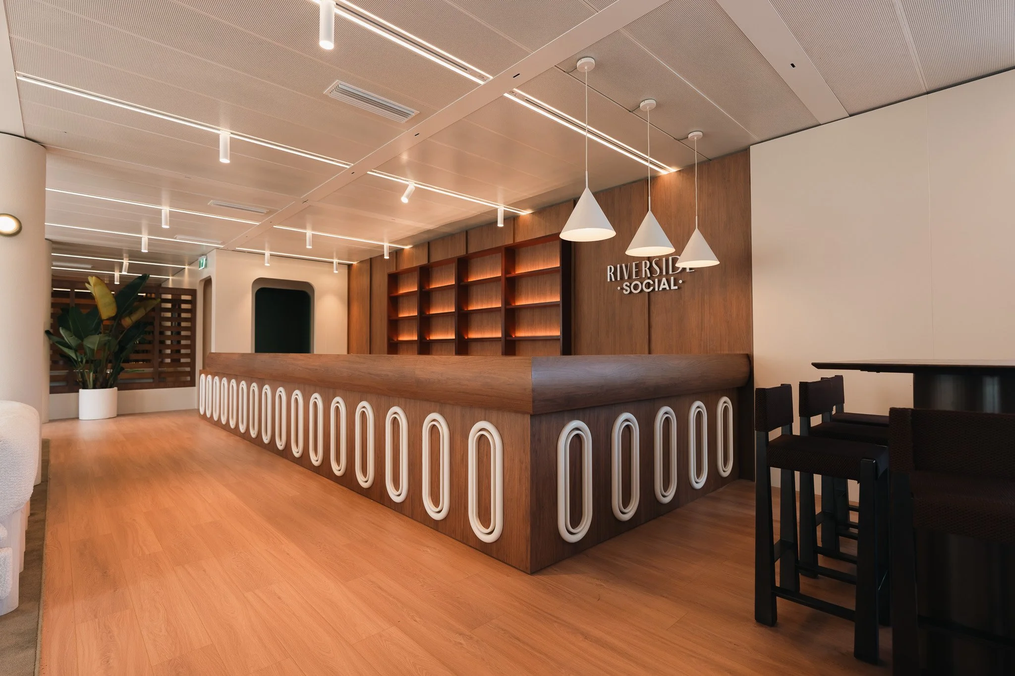 Modern reception area with wooden desk, white decorative accents, hanging cone-shaped light fixtures, wood shelves on the wall, a green plant in a white pot, and the sign 'RIVERSIDE SOCIAL' on the wall.