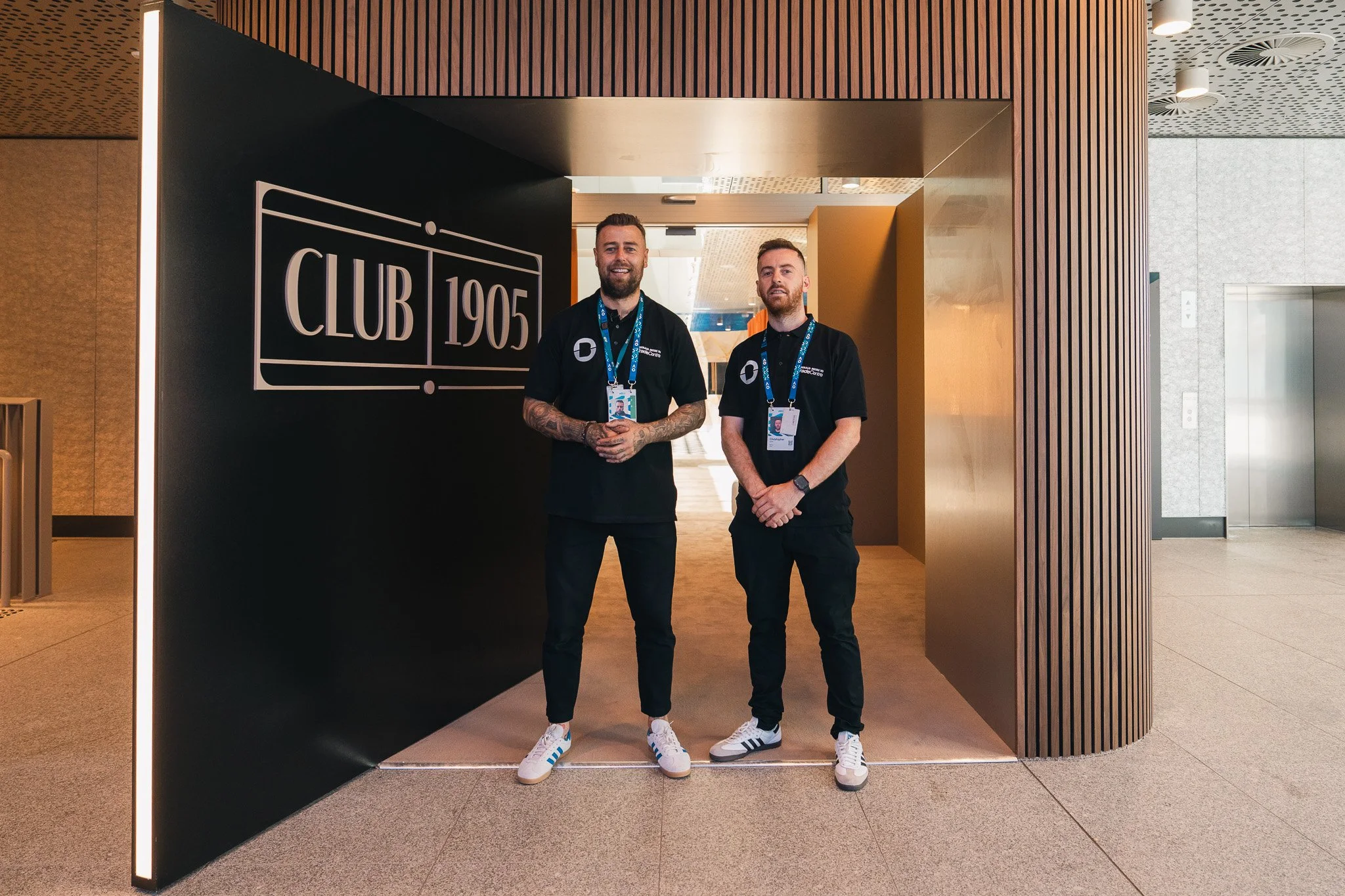 Two men with lanyards standing at the entrance of a modern building with a sign reading 'CLUB 1905', both smiling and dressed in black polo shirts with logos.