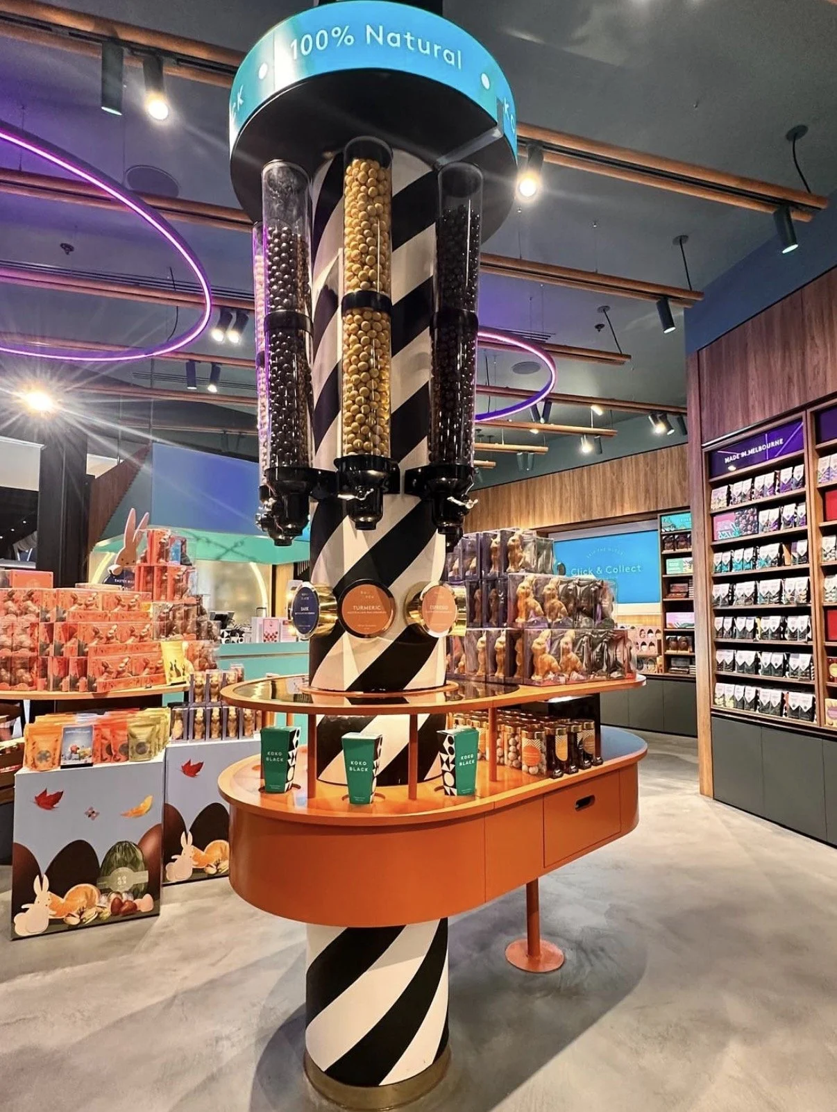 Display inside a store showcasing a spiral display with three vertical tubes filled with colored balls, and a sign on top reading '100% Natural'. Surrounding shelves hold packaged products, likely chocolates or snacks, with colorful packaging and the