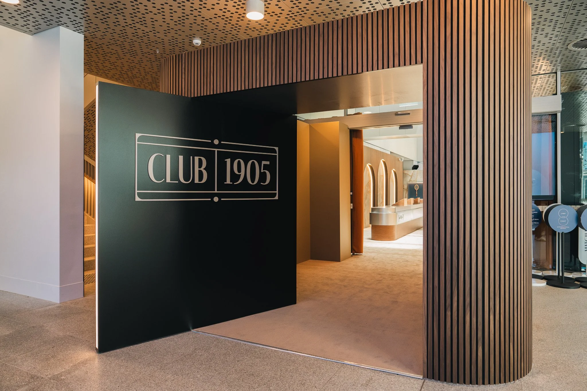 Interior of a modern building with a sign reading 'Club 1905'. Wooden slat wall, curved doorway, and visible interior with lighting fixtures and stairs.