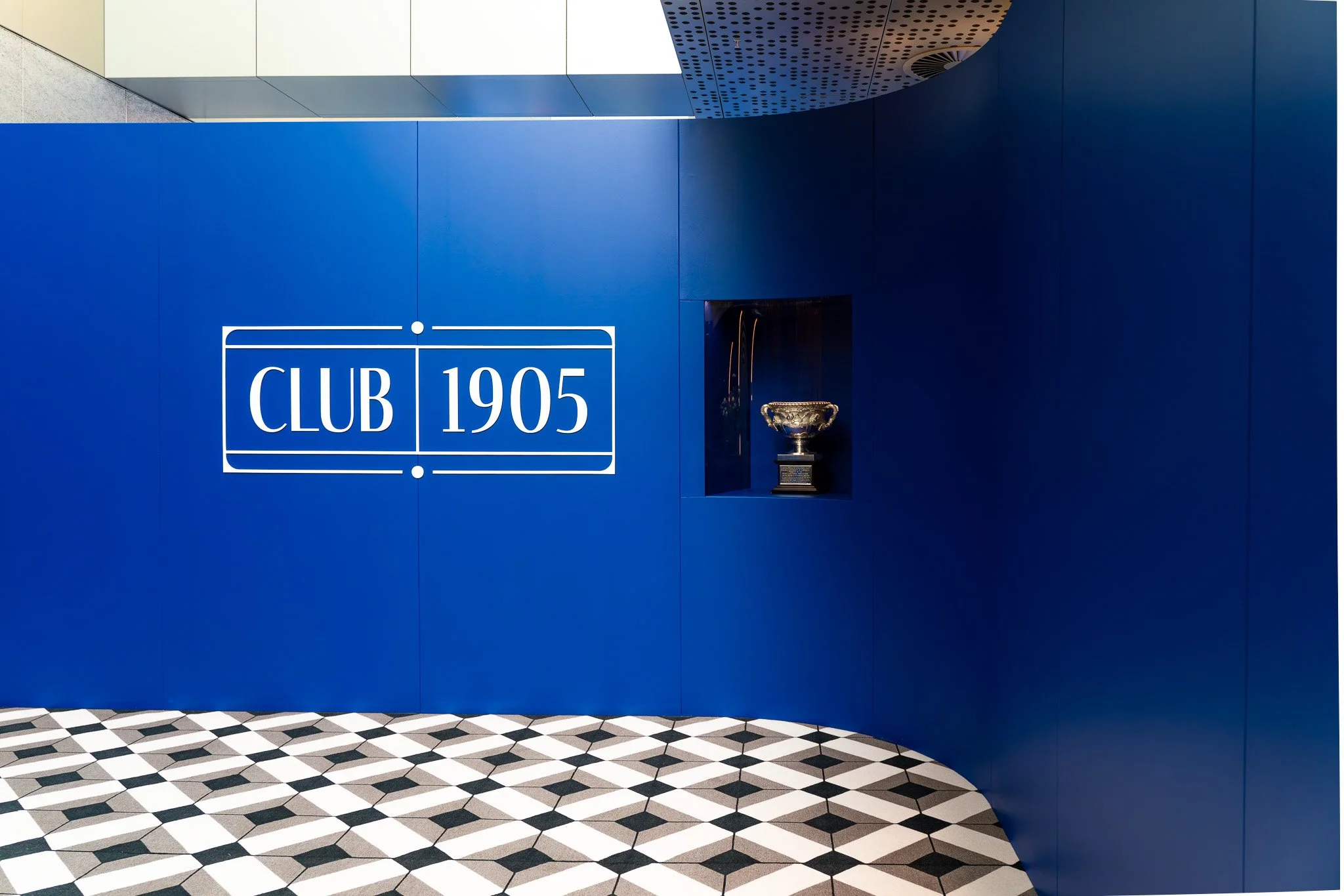 Interior of a modern building with a blue wall featuring a sign that reads 'CLUB 1905' in large white letters, a decorative trophy display, and a black-and-white geometric patterned floor.