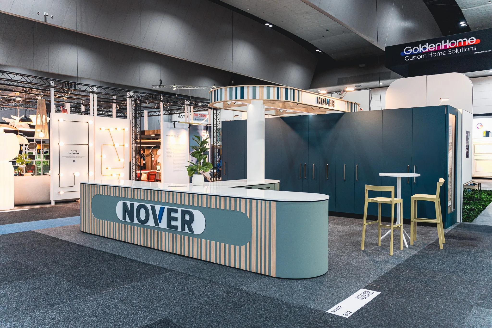Trade show booth for NOVER with a curved reception desk, a small seating area with yellow chairs, and a plant in a white pot. The booth displays modern furniture and lighting, with a sign for GoldenHome in the background.