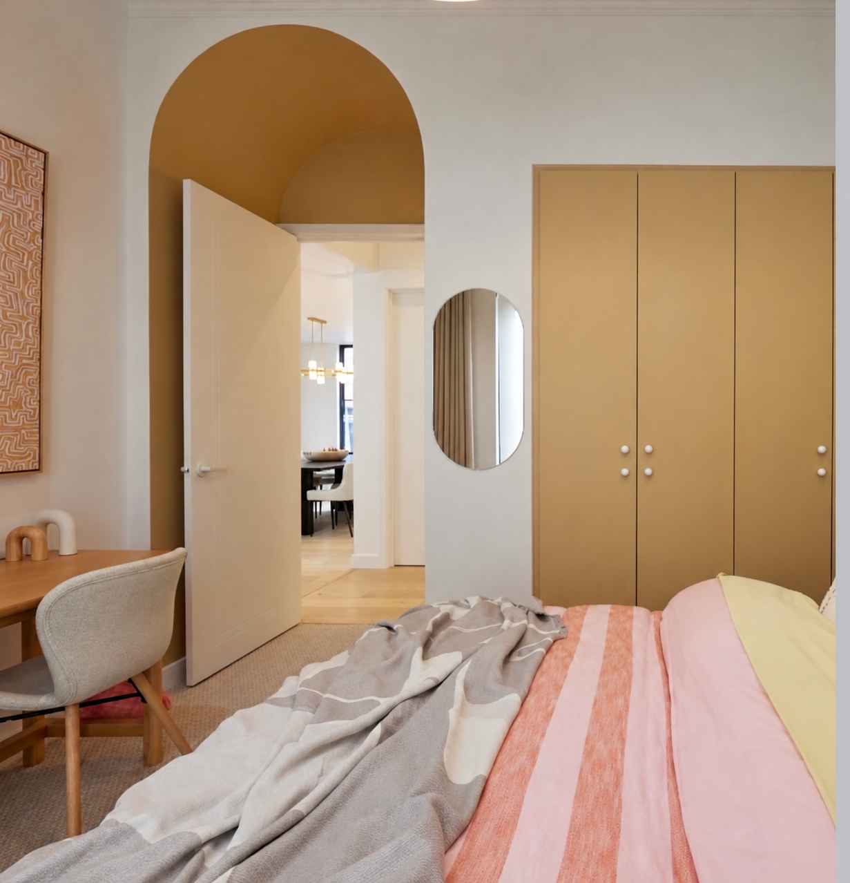 A bedroom with a bed partially covered by a striped blanket, a wooden desk with a beige chair, a mustard-colored closet, a mirror on the wall, and an open doorway leading to a dining area with a table and chairs, in a modern style with neutral tones.