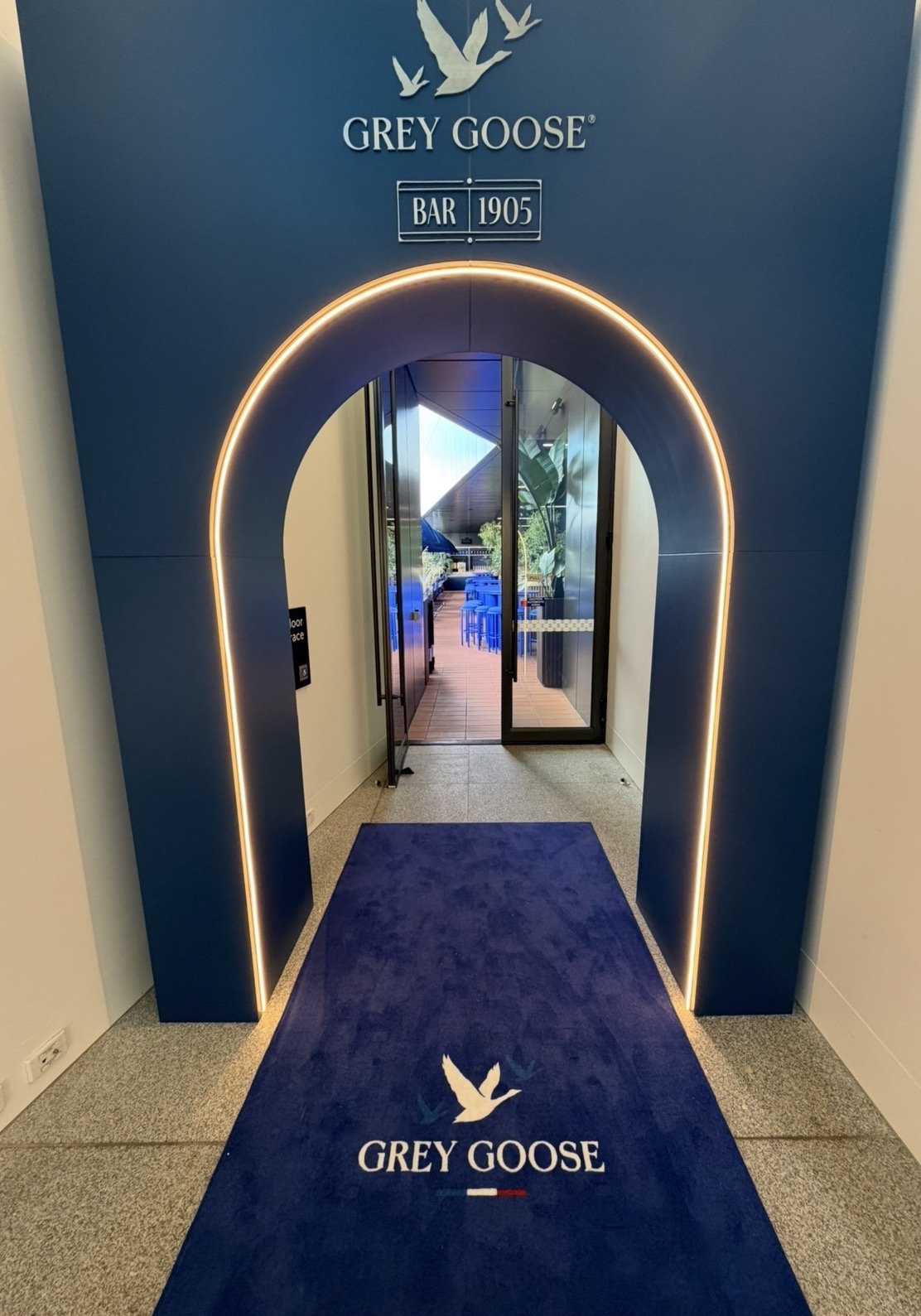 Entrance to Grey Goose bar with illuminated archway, branded logo on a blue carpet and sign, sliding glass doors leading outside to a patio with tables and chairs.
