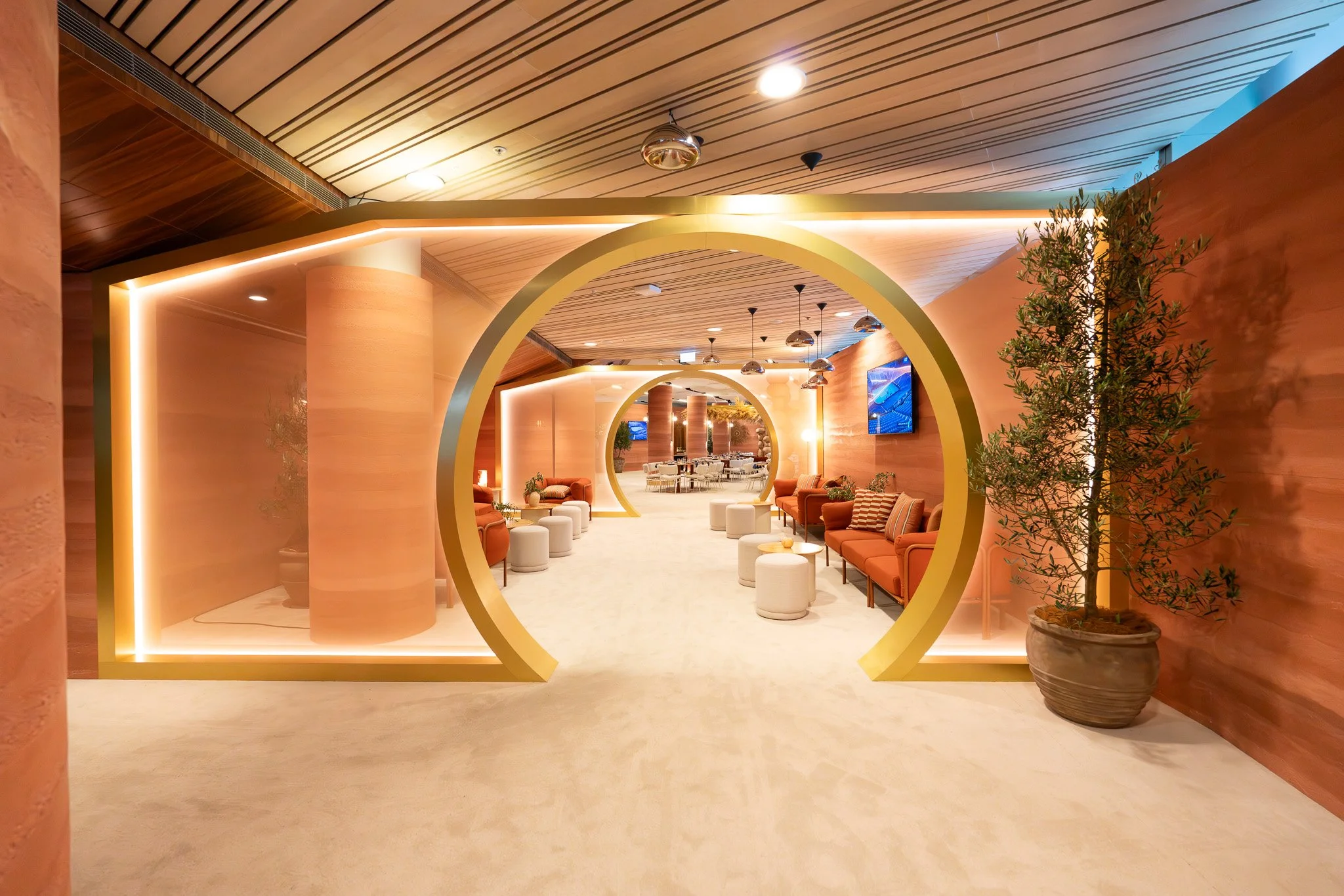 Luxury hotel lobby with warm lighting, rounded archways, orange seating, potted plants, and television screens.