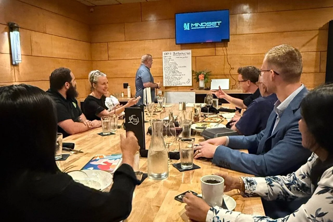 A business meeting taking place around a long wooden table with people engaged in discussion, in a room with wood-paneled walls, a television screen displaying "MINDSET,