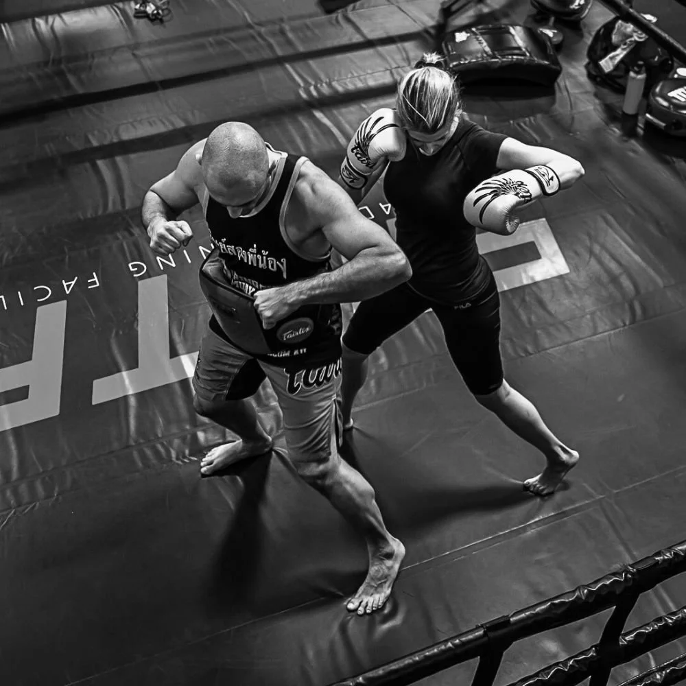Two people practicing martial arts in a boxing ring, one sparring partner is a man with a shaved head wearing a black tank top and gray shorts, and the other is a woman with blonde hair tied back, wearing a black shirt, black shorts, and yellow boxing gloves. They are engaged in a close combat stance.