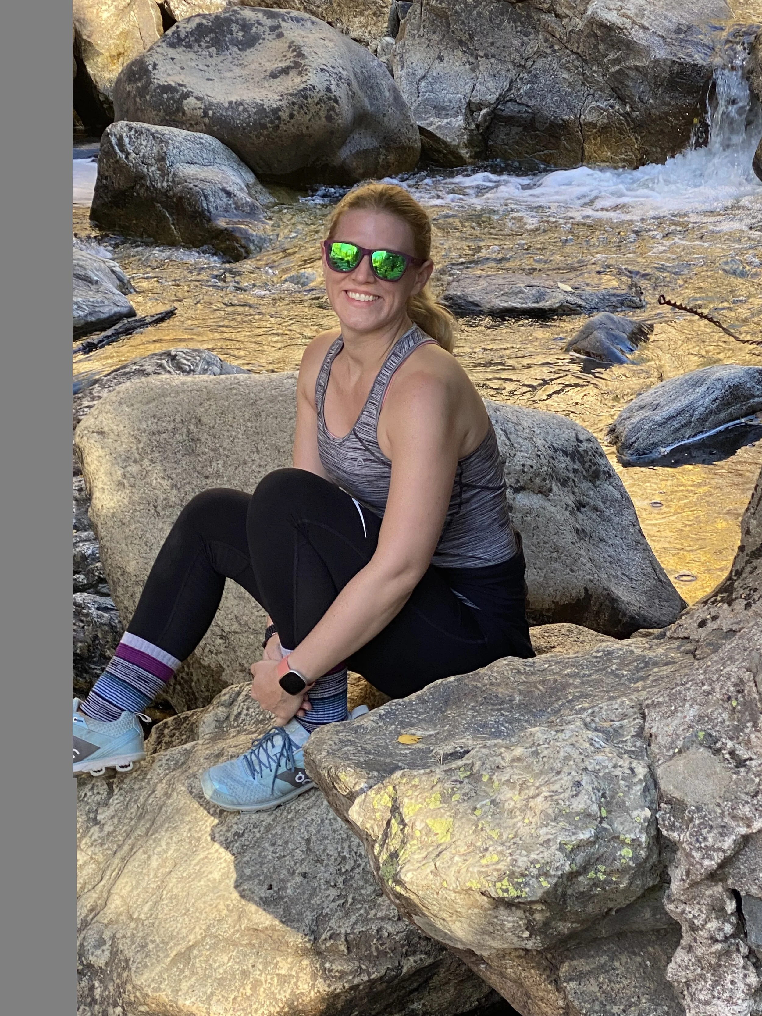 A woman wearing sunglasses, a gray athletic tank top, black leggings, blue running shoes, and a fitness tracker is sitting on a large rock near a small waterfall in a rocky outdoor setting, smiling at the camera.