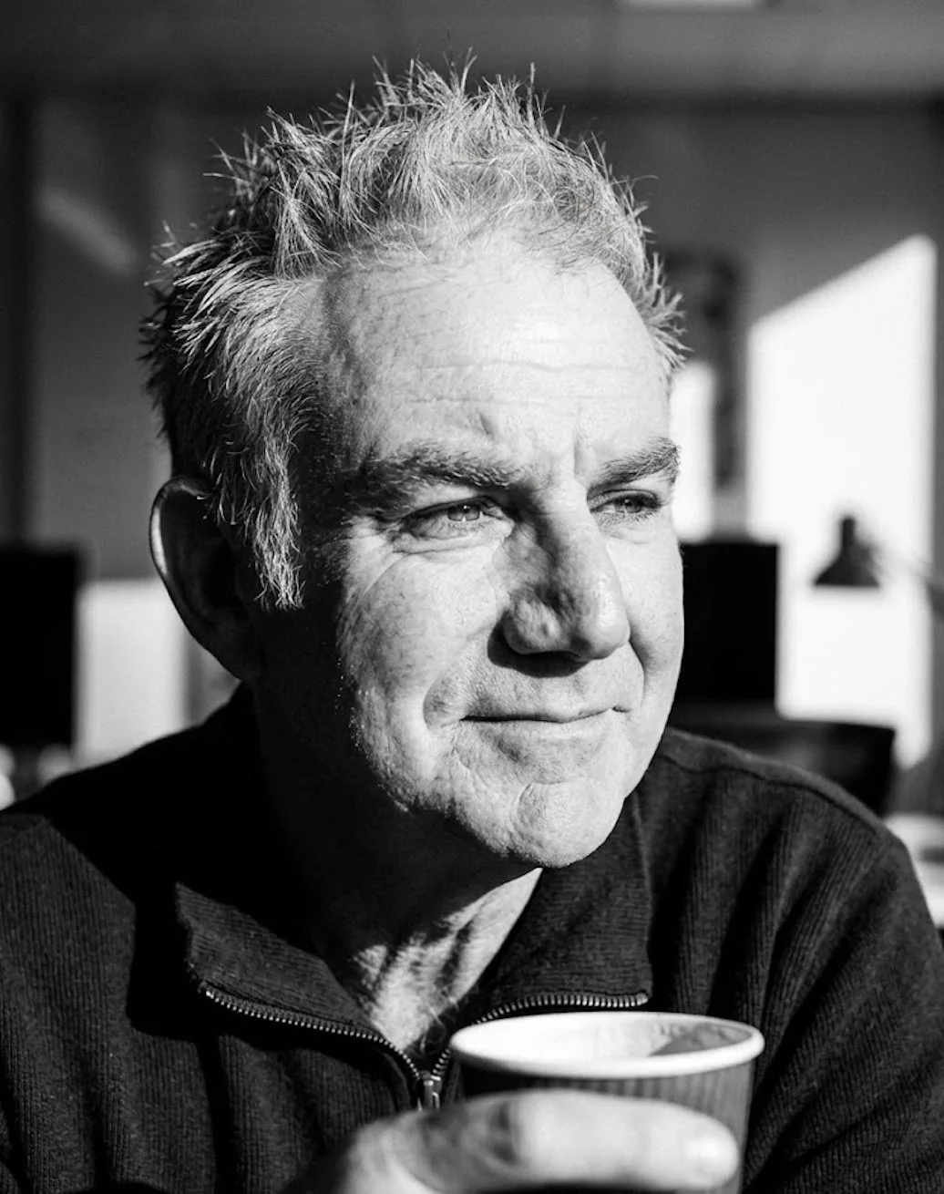 A black and white photo of an older man with gray hair, smiling slightly while holding a cup, sitting indoors with a blurred background.
